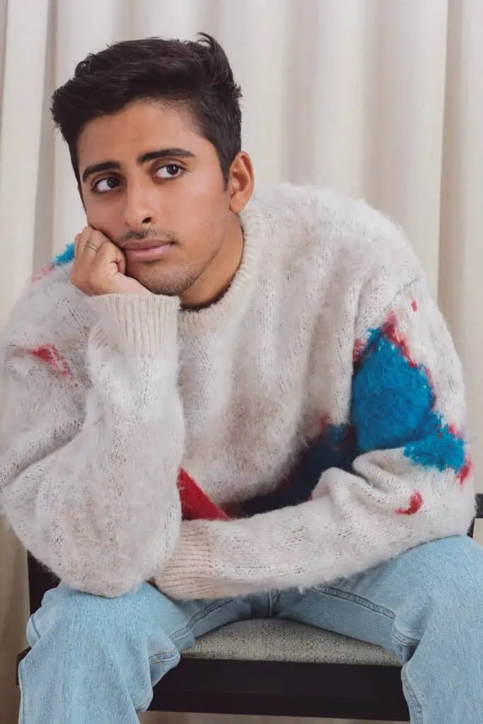 A person with short dark hair, resembling Karan Brar, wearing a fuzzy cream sweater with red and blue patches and light blue jeans, sits on a chair resting their chin on their hand, gazing thoughtfully to the side. Cream curtains are in the background.