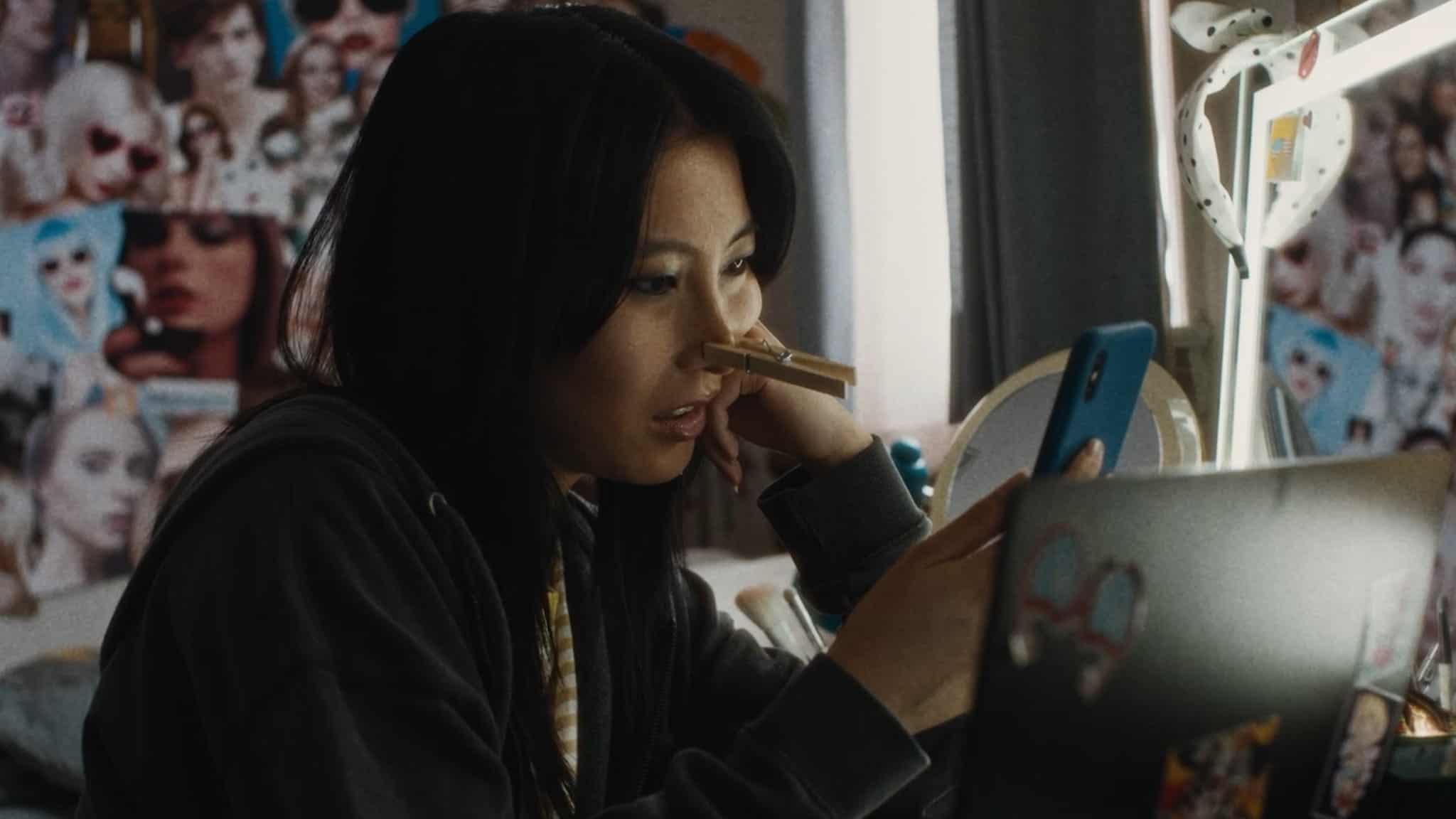 A young woman sits at a desk, looking at her phone with a clothespin clipped to her nose. She wears a dark hoodie, and behind her is a wall covered with photos and posters.