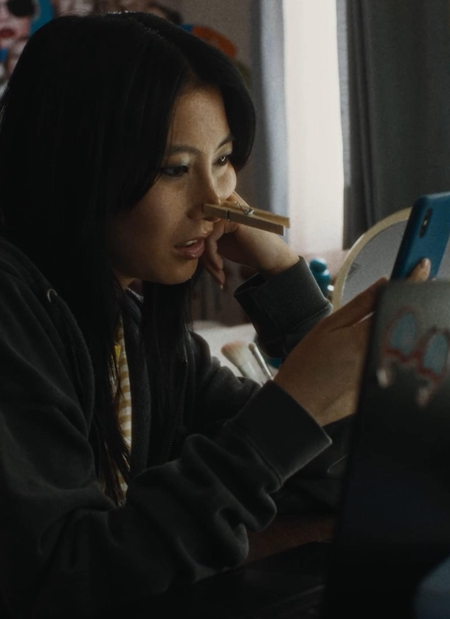 A young woman sits at a desk, looking at her phone with a clothespin clipped to her nose. She wears a dark hoodie, and behind her is a wall covered with photos and posters.