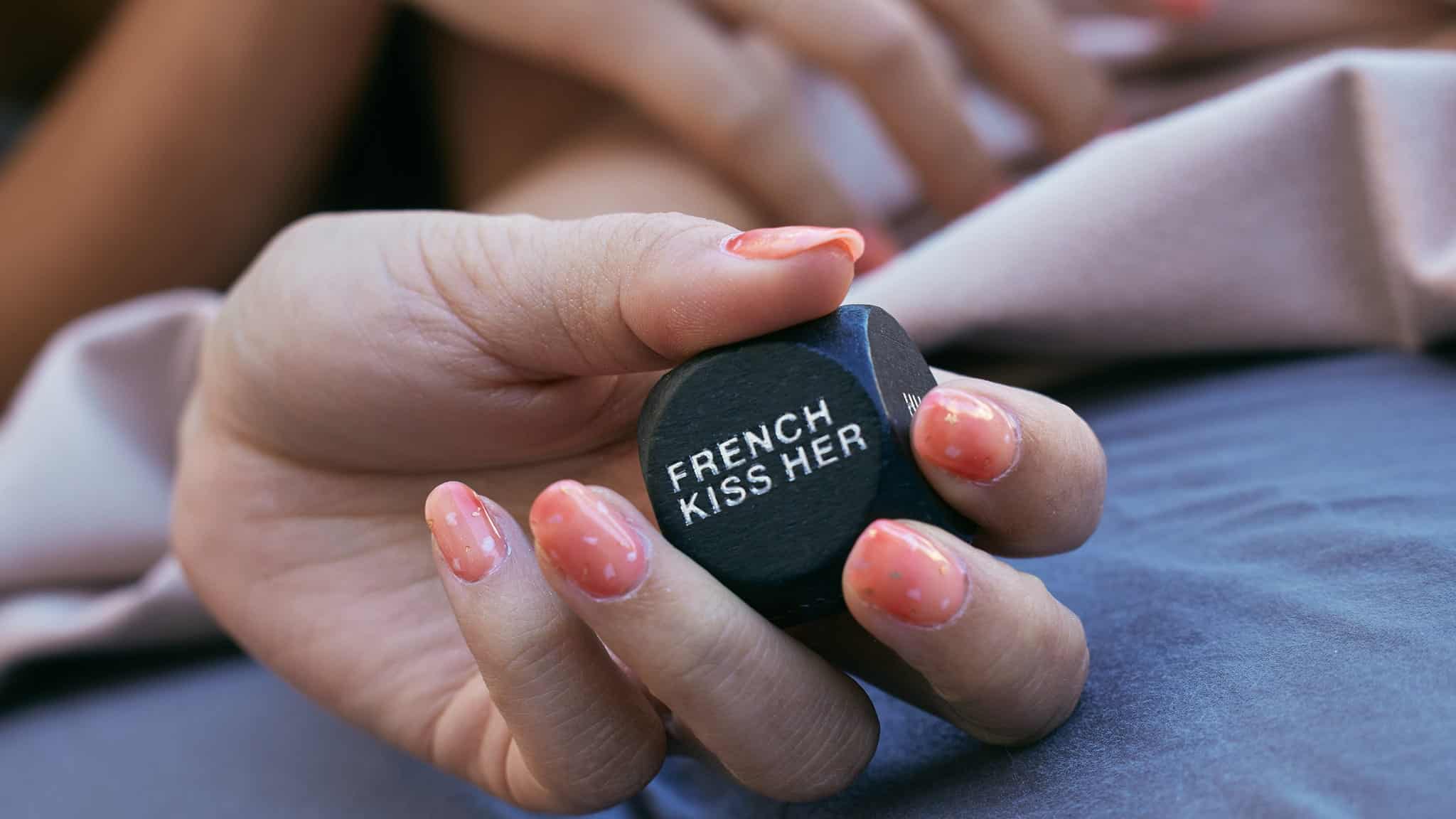 A hand with pink, speckled nails holds a black die with the words FRENCH KISS HER written on it. A blurred background shows another hand and part of a bed with sheets.