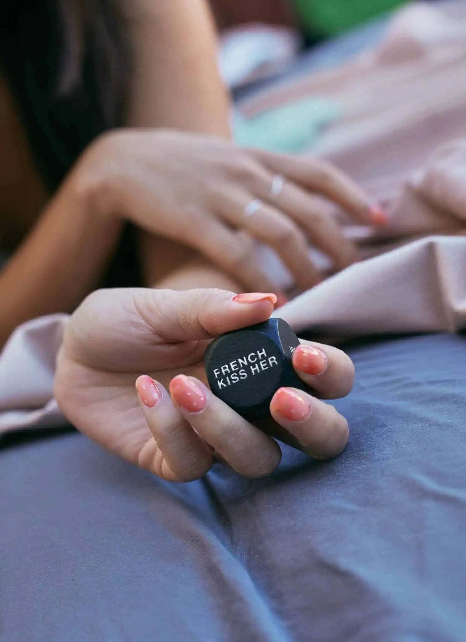 A hand with pink, speckled nails holds a black die with the words FRENCH KISS HER written on it. A blurred background shows another hand and part of a bed with sheets.
