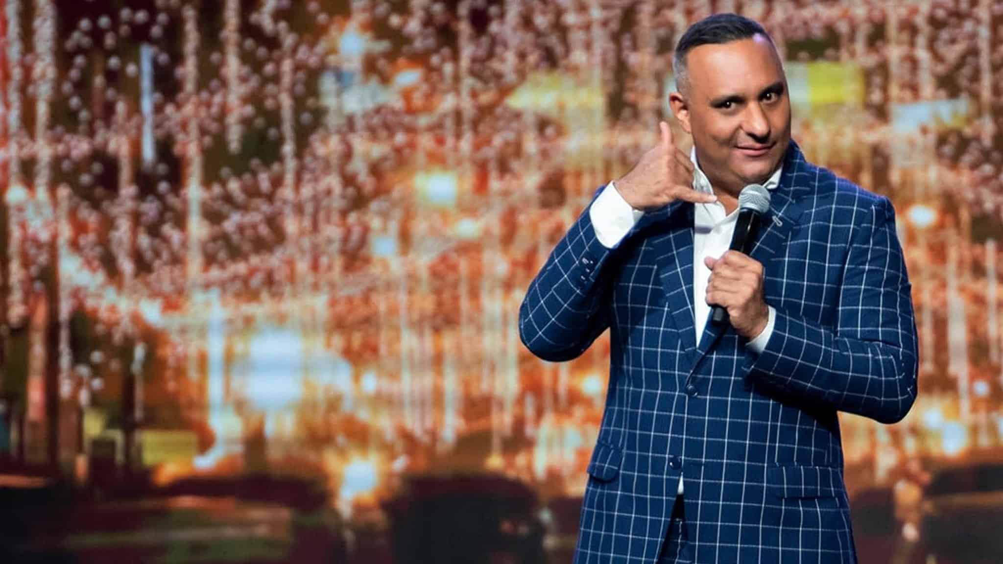 A comedian in a blue checkered suit holds a microphone and makes a hand gesture resembling a phone while performing on stage with twinkling lights in the background.