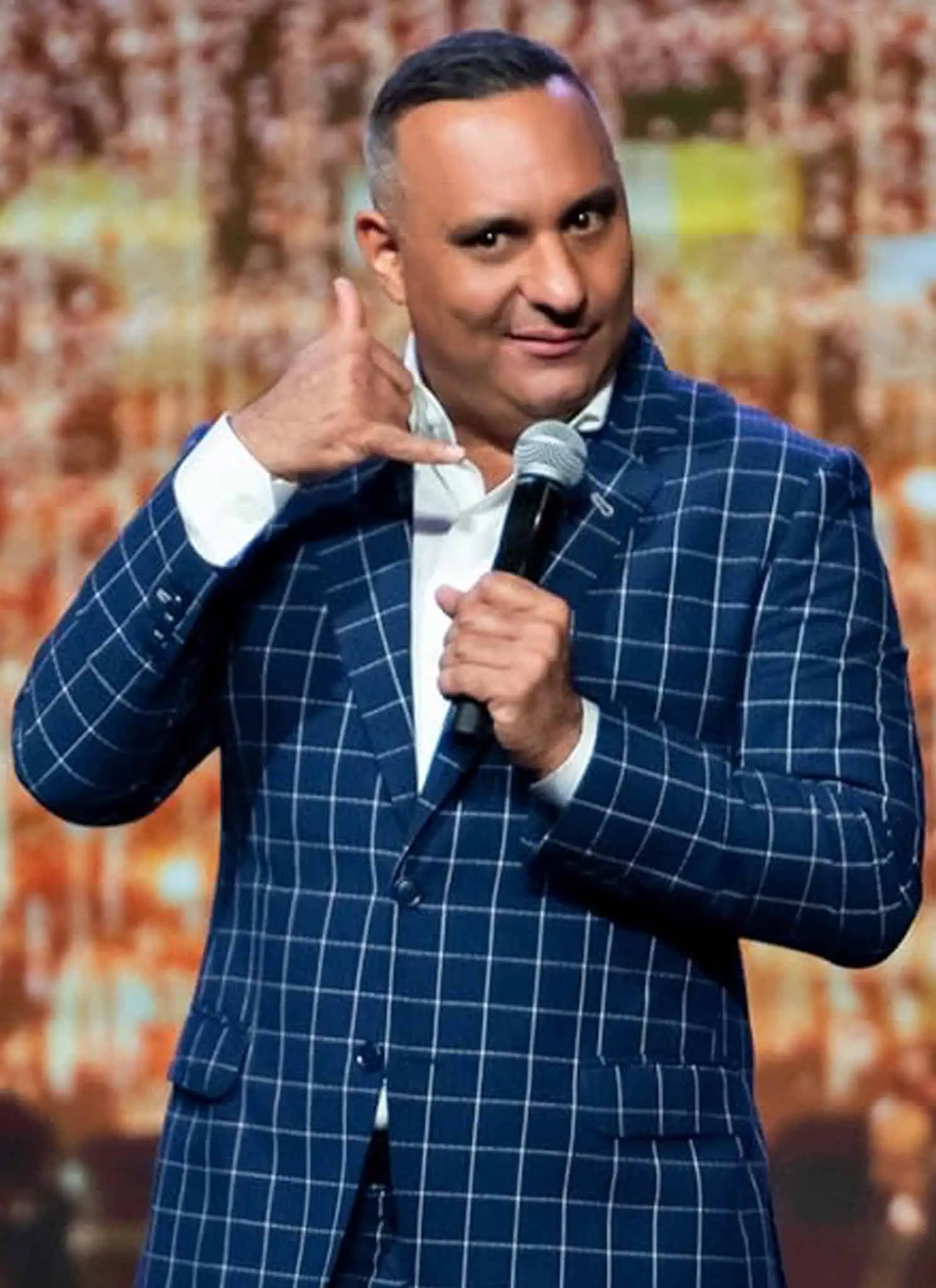 A comedian in a blue checkered suit holds a microphone and makes a hand gesture resembling a phone while performing on stage with twinkling lights in the background.
