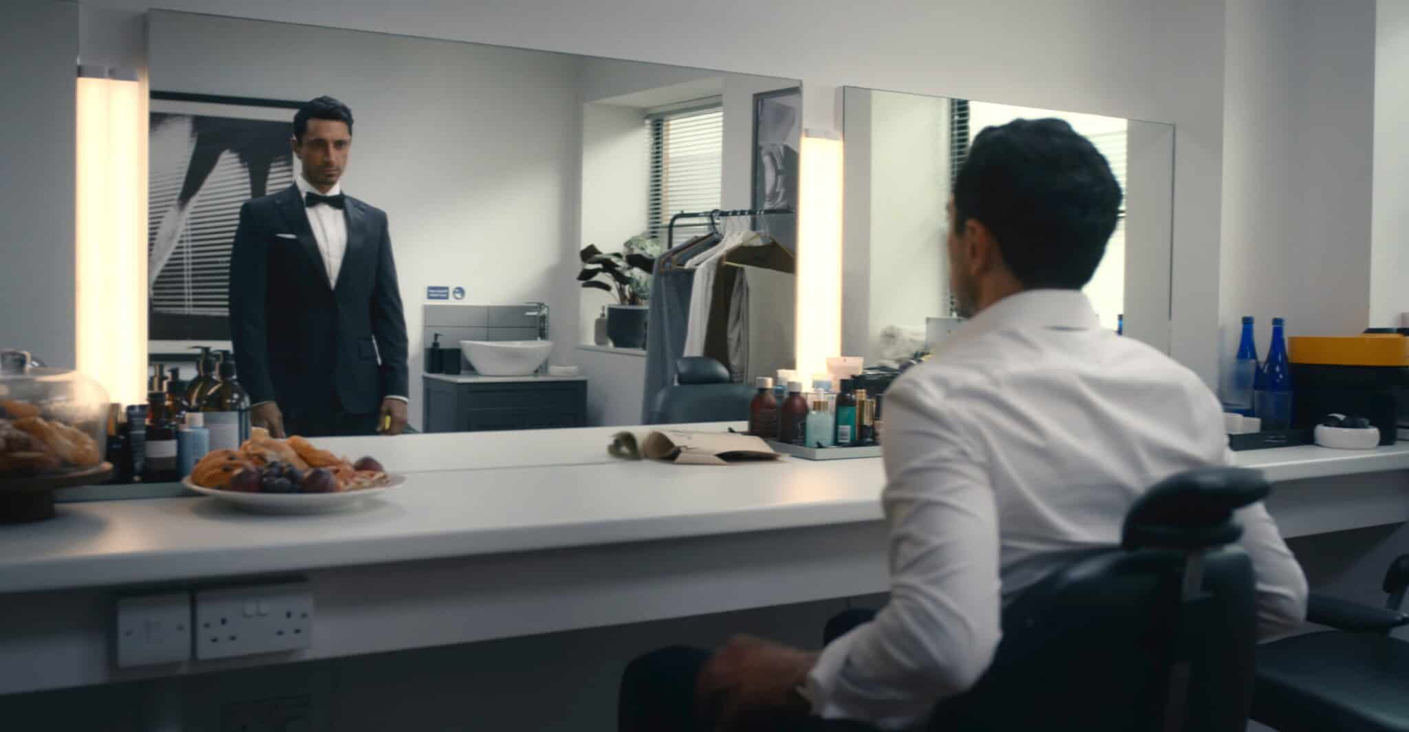 A man in a white shirt sits at a dressing room table, looking into a mirror where his reflection appears dressed in a formal suit and bow tie. The room has cosmetics, snacks, and clothes hanging in the background.