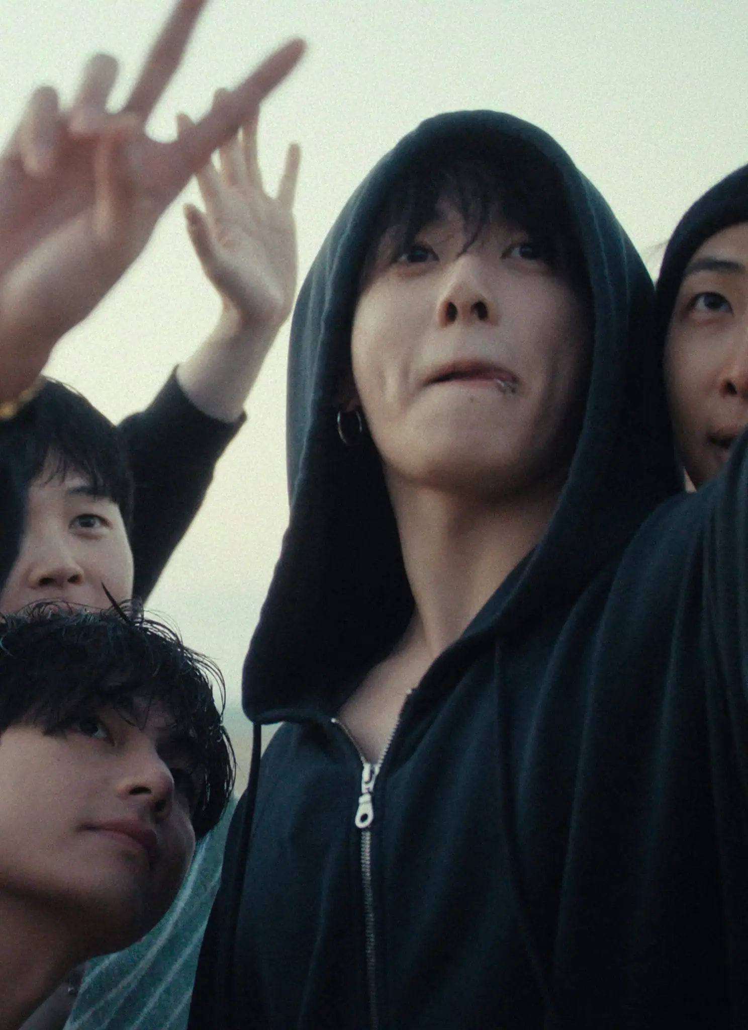 A group of young men posing together outdoors, some making peace signs and smiling. They appear happy and are dressed casually in hoodies, beanies, and sunglasses. The background is a bright, clear sky.