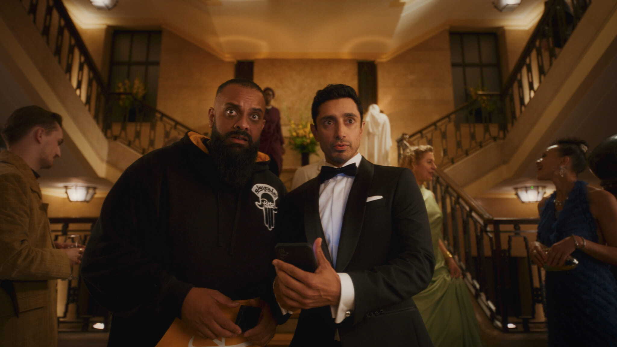 Two men stand at the center of a grand staircase; one wears a black hoodie and looks surprised, while the other, in a tuxedo, holds a phone. People in formal attire mingle in the background.
