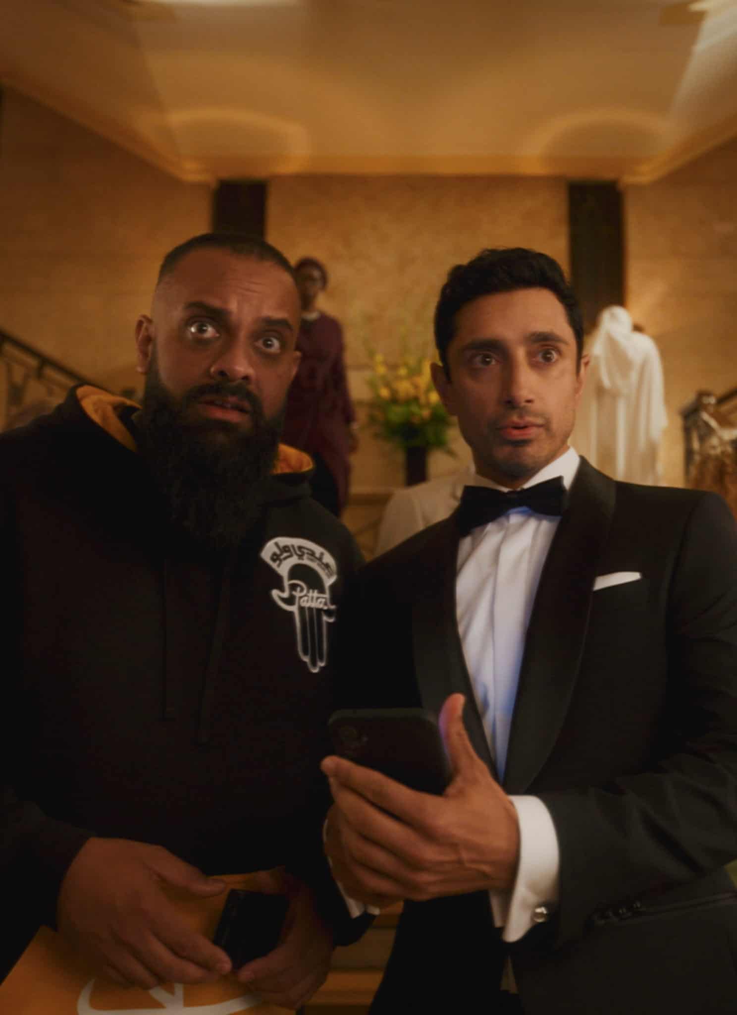 Two men stand at the center of a grand staircase; one wears a black hoodie and looks surprised, while the other, in a tuxedo, holds a phone. People in formal attire mingle in the background.