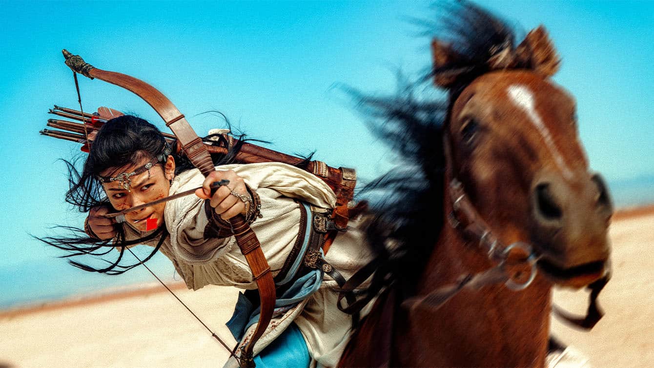 A woman in traditional attire rides a galloping horse, aiming a drawn bow and arrow with intense focus against a bright blue sky and desert landscape. Her hair and the horse’s mane flow dramatically in the wind.