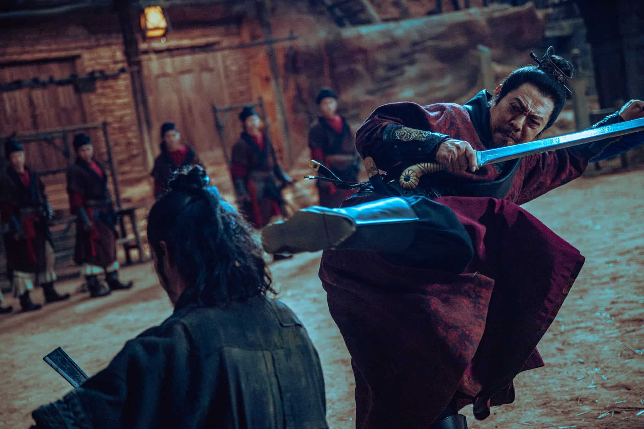A man in traditional Asian attire kicks another man while wielding a sword in a dramatic indoor fight scene, with several onlookers dressed in red robes standing in the background.