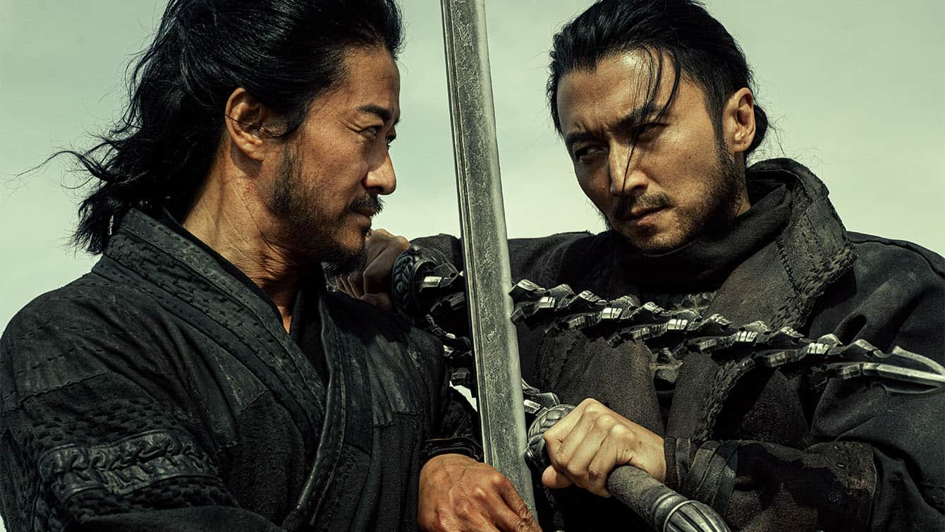 Two men in dark, traditional warrior clothing face each other intently. One holds a sword with a chained blade, pressing it against the others arm as they engage in a tense standoff against a cloudy sky.