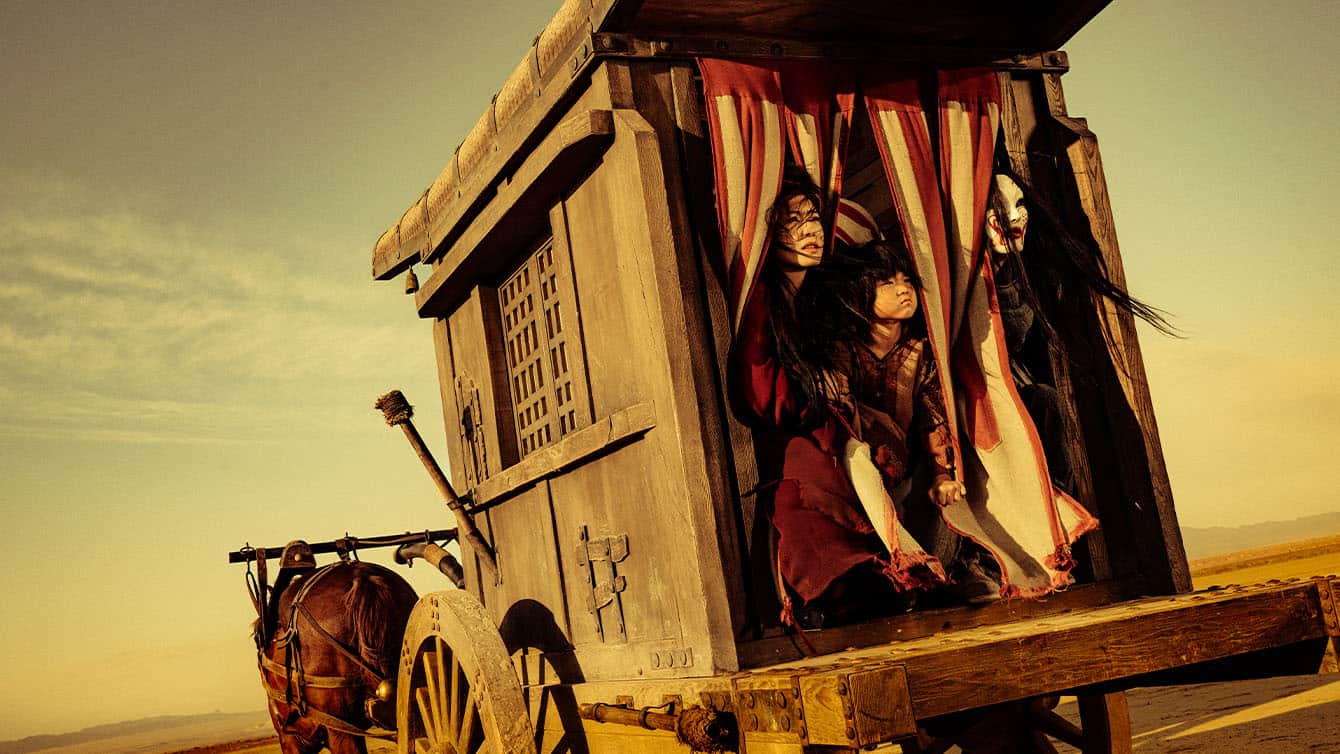 Two people in dramatic makeup and costumes peer out from the red-striped curtains of a wooden, old-fashioned wagon, which is being pulled by a horse across a dry, open landscape under a golden sky.