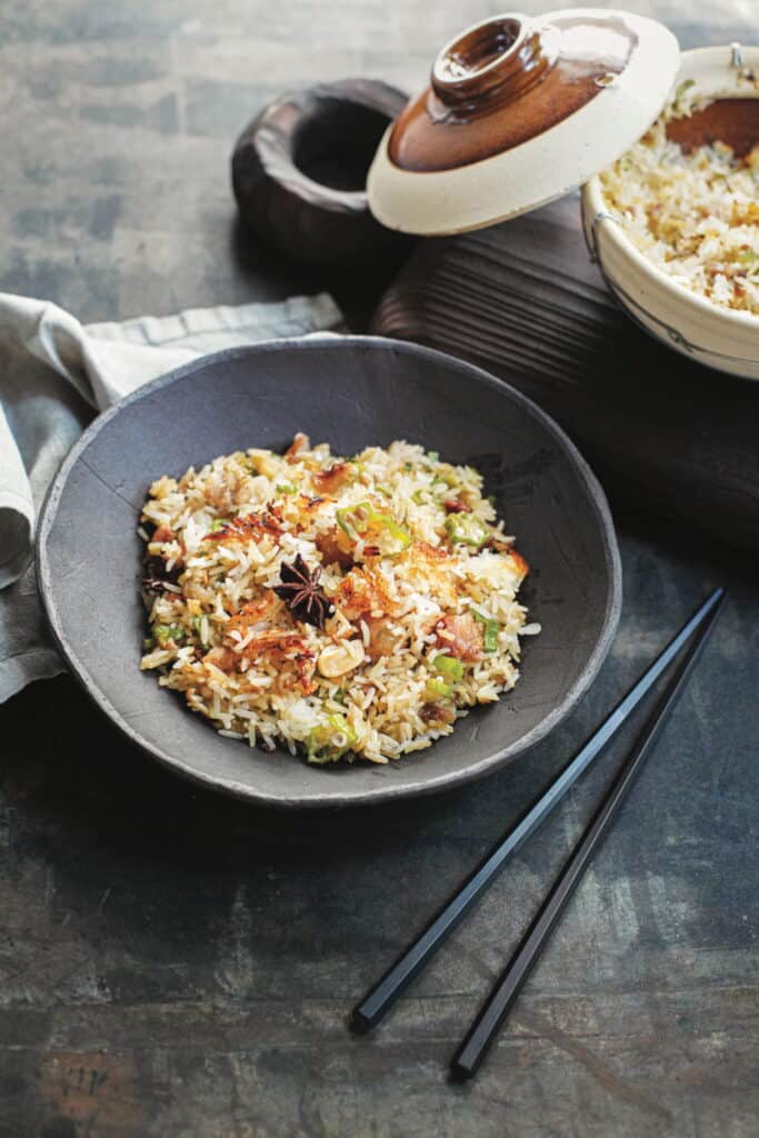 A bowl of fried rice garnished with vegetables and a star anise, placed on a dark surface with chopsticks beside it and a pot of rice in the background.