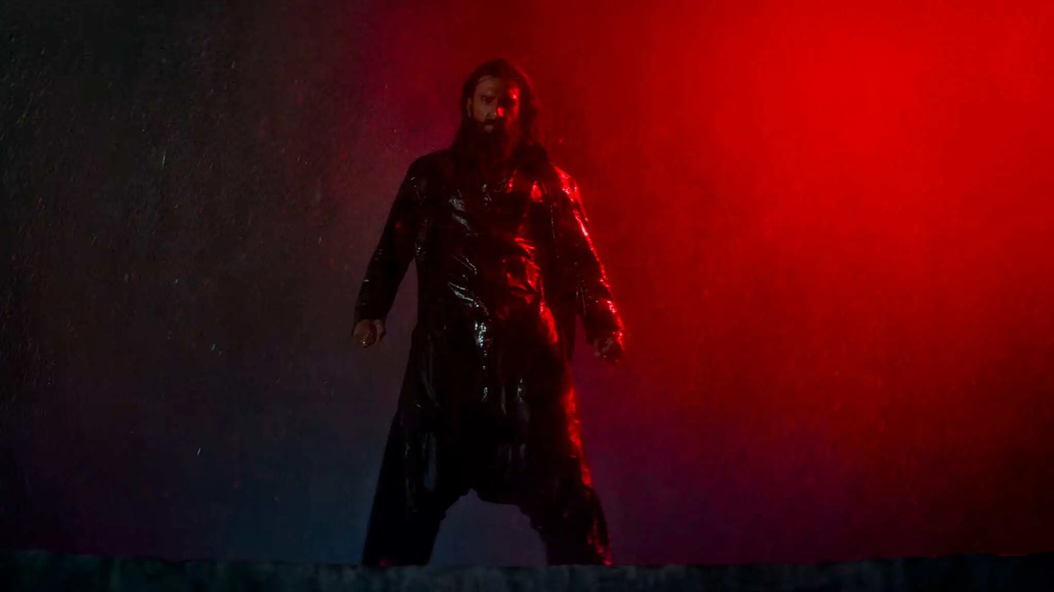 A person with long hair and a beard stands in the rain, wearing a shiny black coat, dramatically lit by a red light on the right side of the image.