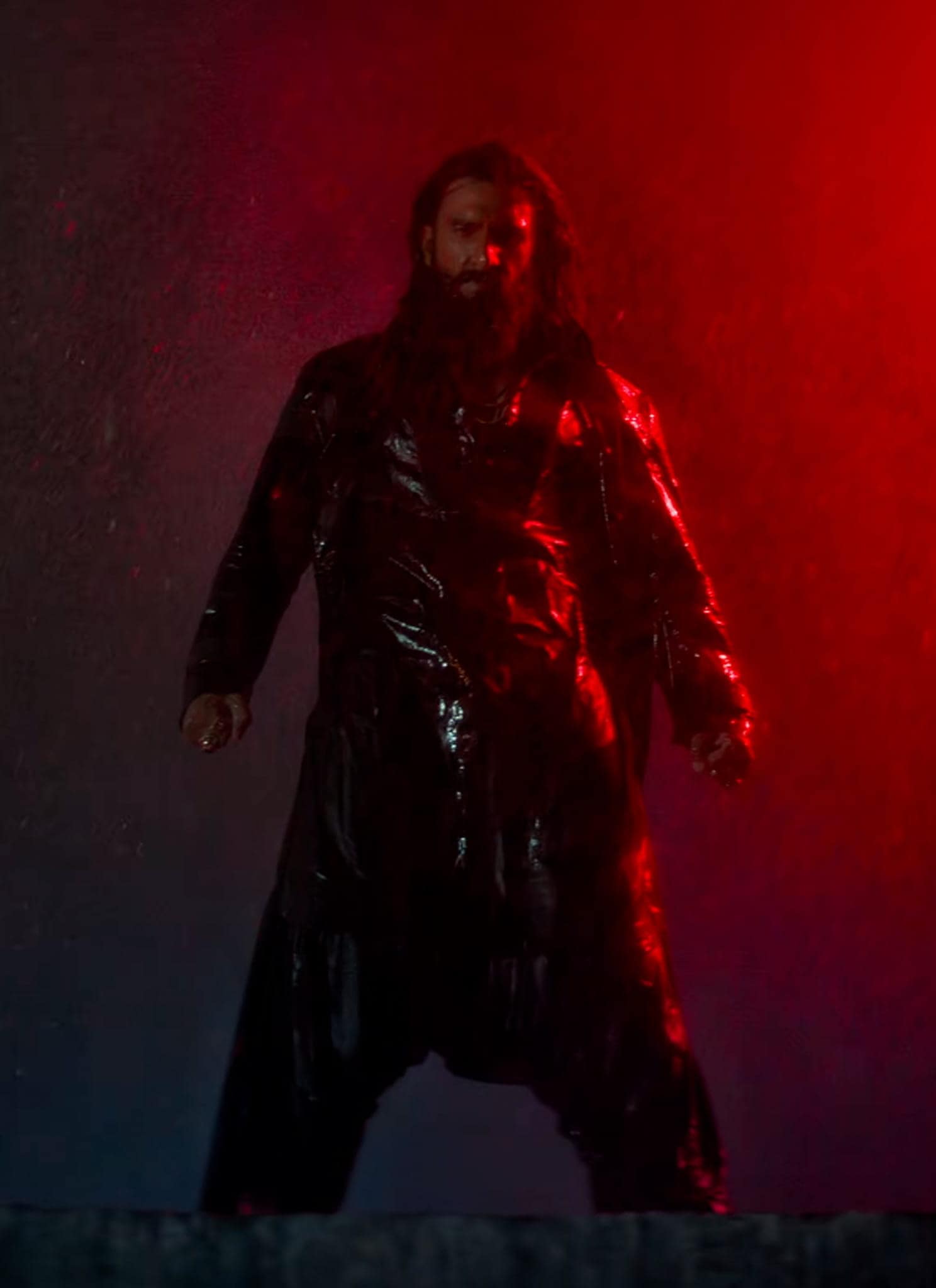 A person with long hair and a beard stands in the rain, wearing a shiny black coat, dramatically lit by a red light on the right side of the image.