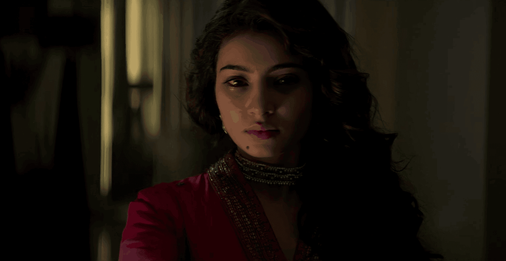 A woman with long, wavy hair and a serious expression wears a red traditional outfit with a decorative collar, standing in soft, dim lighting with a blurred background.