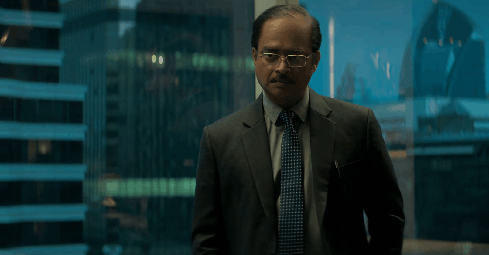 A man in a suit and tie stands in front of a large window overlooking tall city buildings, looking serious and thoughtful.