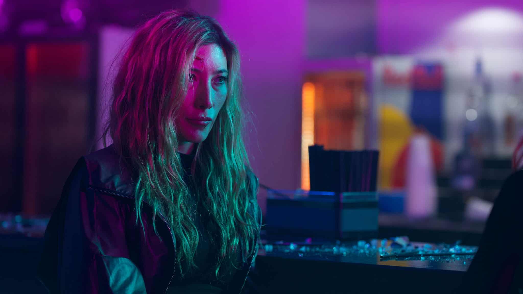 A woman with long, wavy blonde hair sits at a bar bathed in vibrant pink and teal lighting, looking serious and thoughtful. The background is blurred with neon lights and bar items visible.