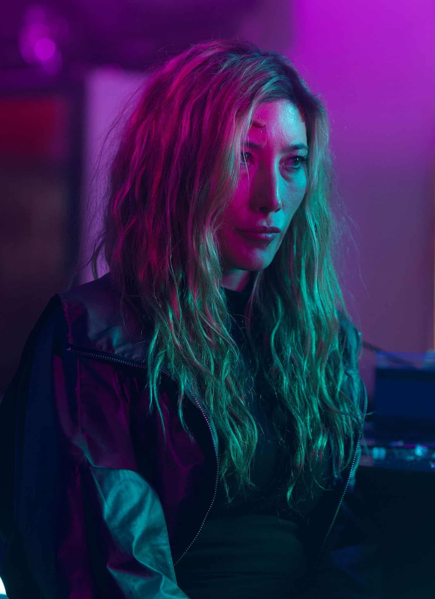 A woman with long, wavy blonde hair sits at a bar bathed in vibrant pink and teal lighting, looking serious and thoughtful. The background is blurred with neon lights and bar items visible.