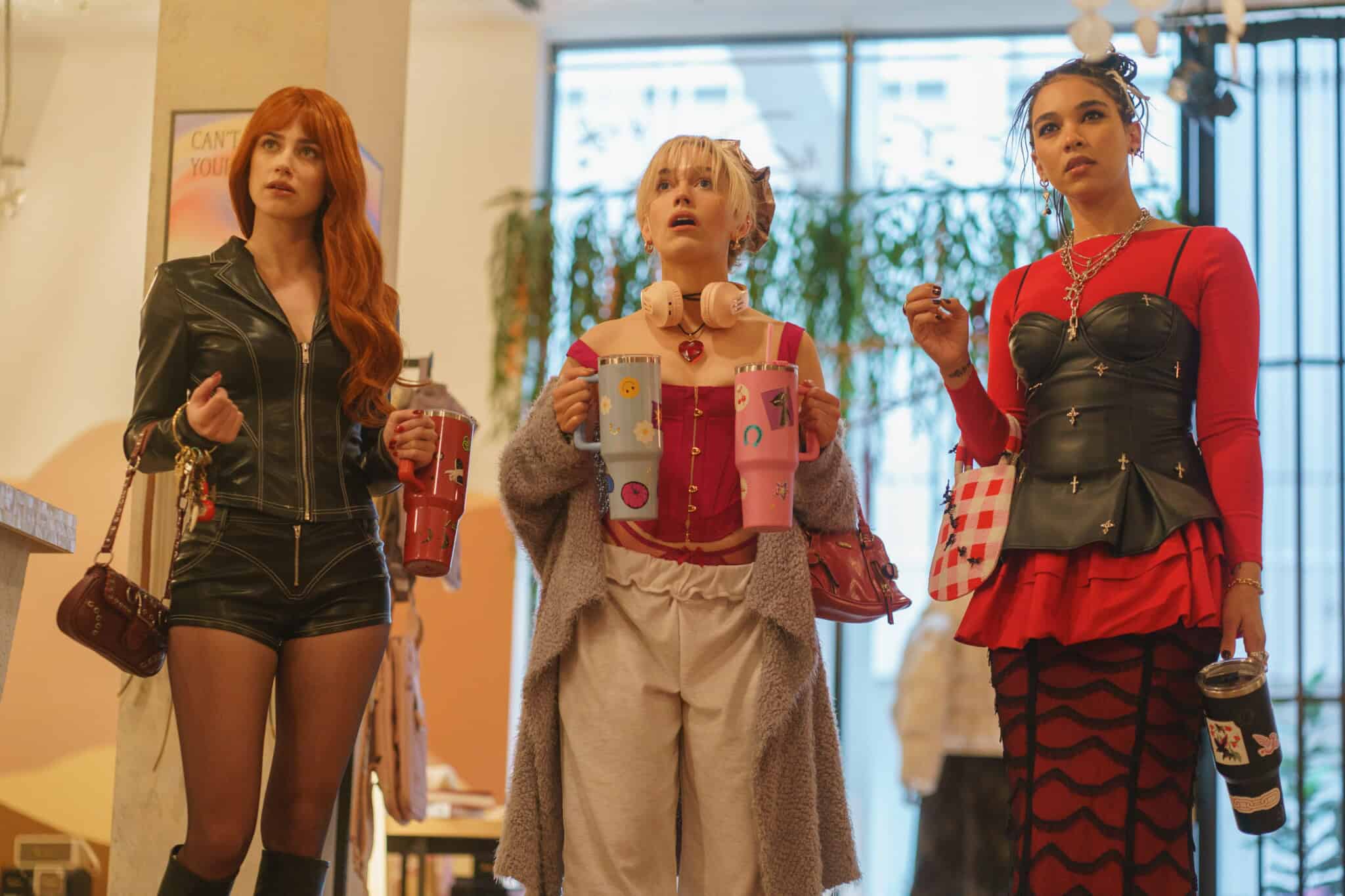 Three young women in fashionable outfits stand indoors, each holding a drink. They look surprised or amazed, facing the same direction. The background shows plants, large windows, and warm lighting.