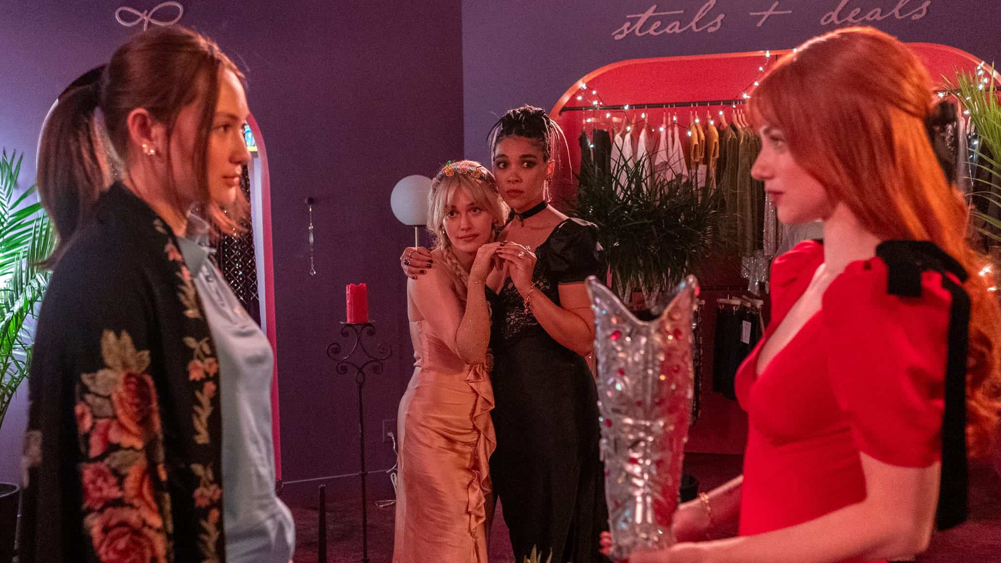 Four women stand in a colorful, dimly-lit room. Two face each other in the foreground, one in a red dress holding a silver vase, while two others, looking concerned, stand close together in the background.