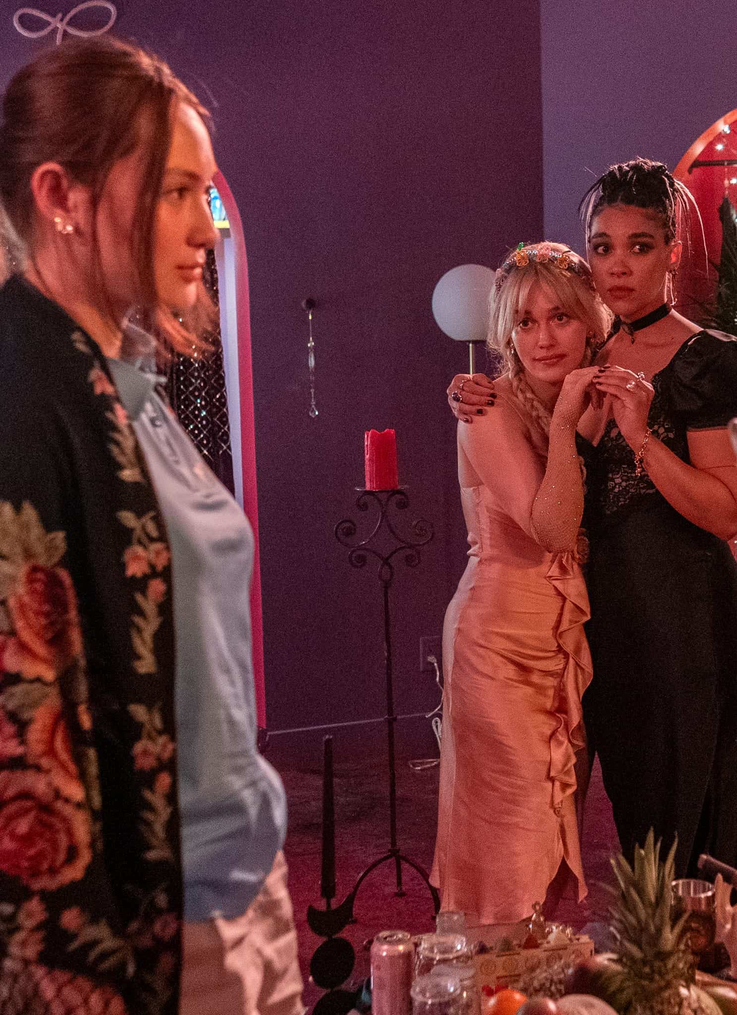 Four women stand in a colorful, dimly-lit room. Two face each other in the foreground, one in a red dress holding a silver vase, while two others, looking concerned, stand close together in the background.