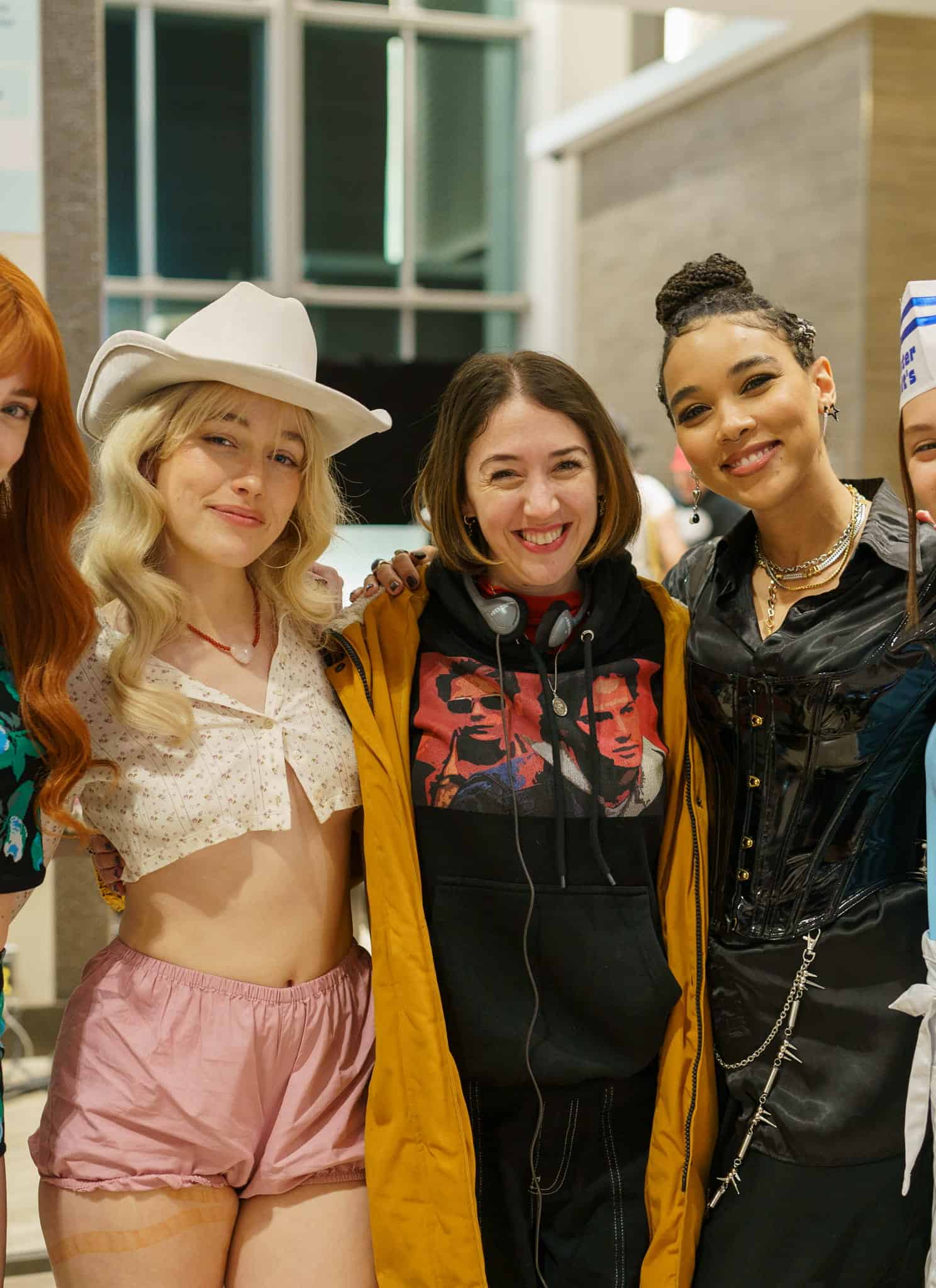 Five young women stand closely together smiling indoors; four wear costumes, including floral shorts, pink shorts with a cowboy hat, a black outfit with chains, and a blue waitress uniform, while one wears a hoodie and jacket.