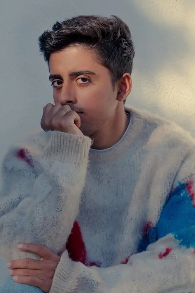 A person with short dark hair, resembling Karan Brar, wears a cream-colored sweater with red and blue patches, sits against a light wall, resting their chin on their hand and looking thoughtfully at the camera.