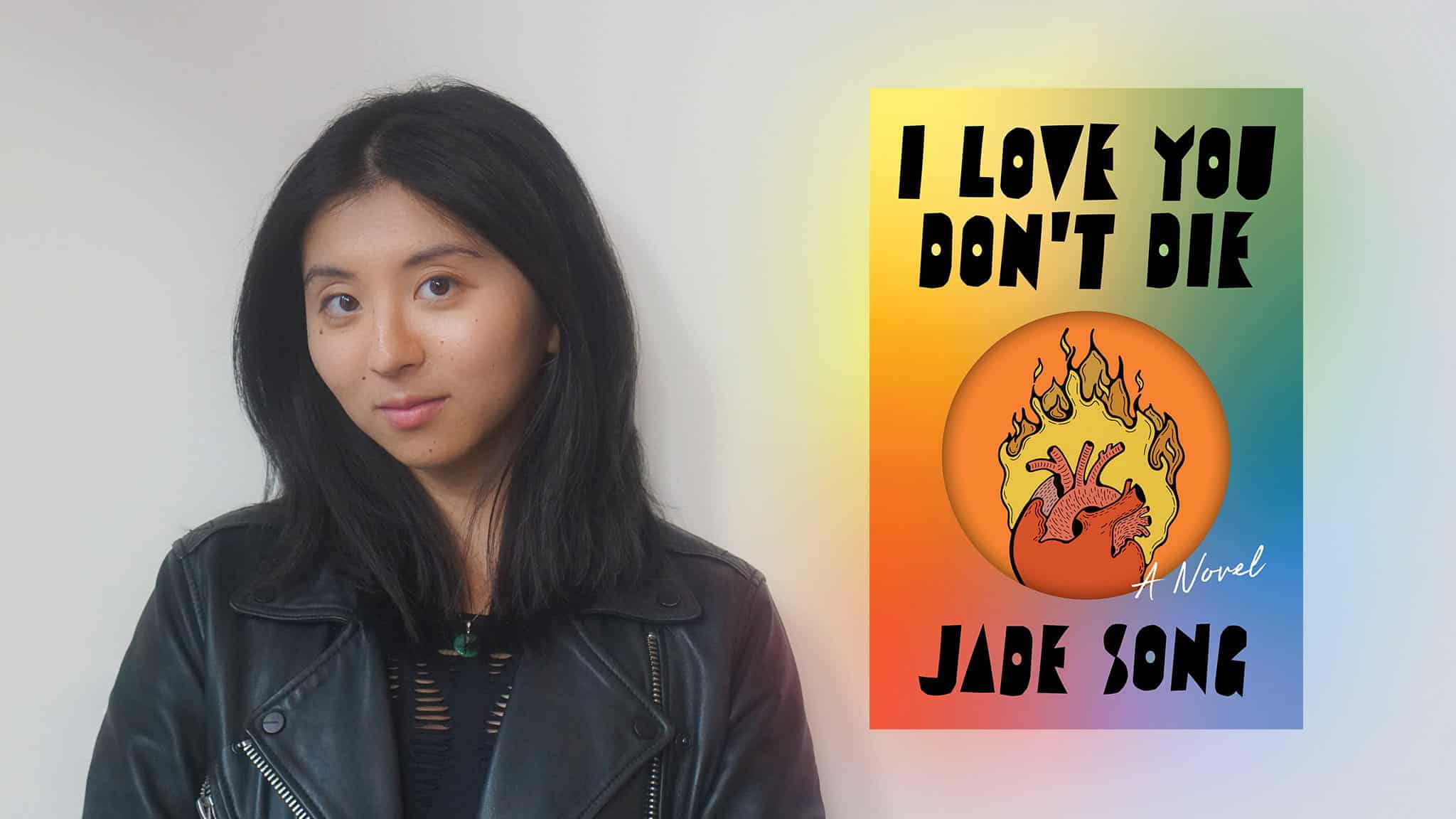 A woman with long black hair in a black leather jacket stands next to the cover of a book titled I Love You Don’t Die by Jade Song, featuring a flaming anatomical heart on a rainbow gradient background.