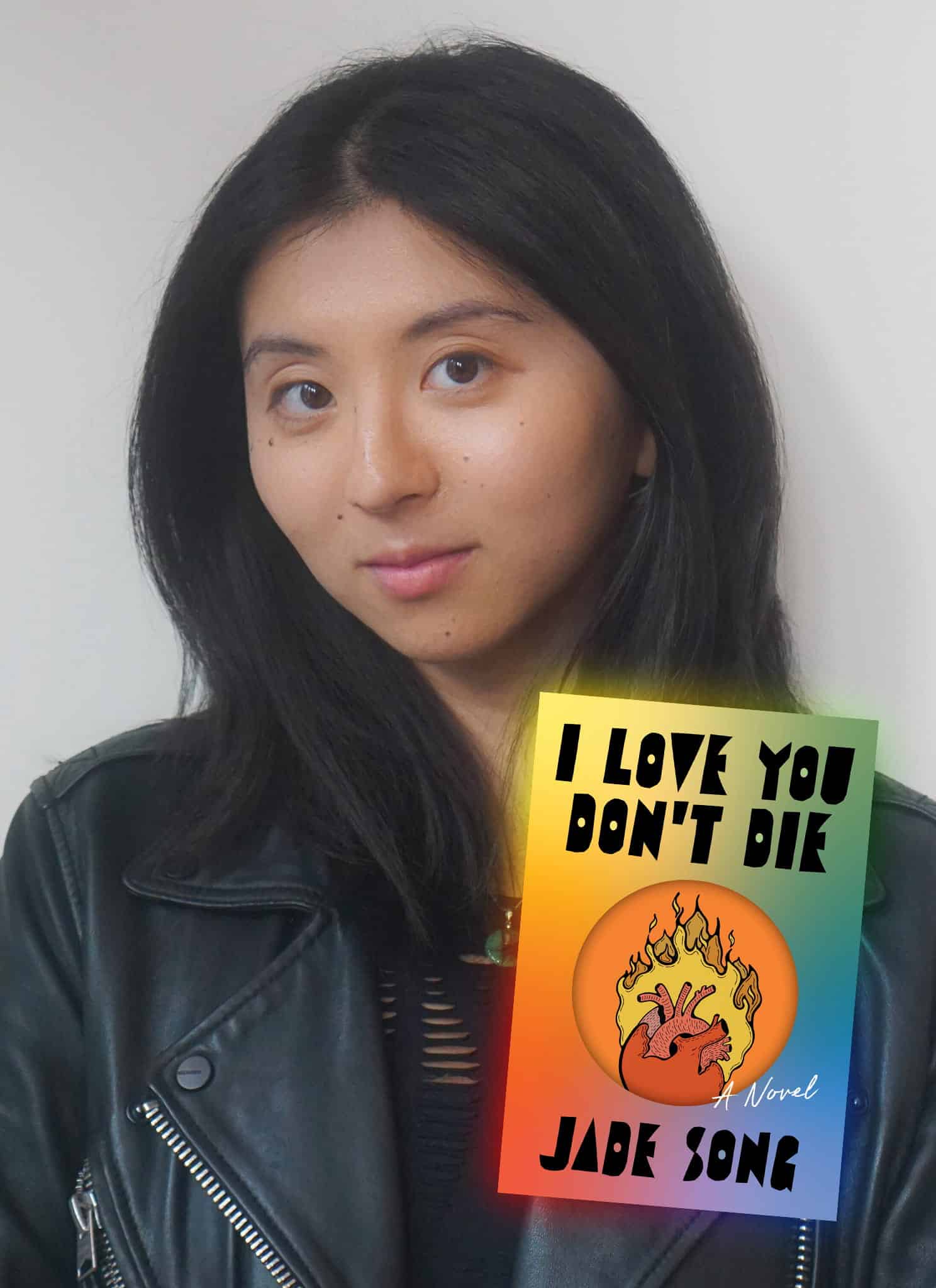 A woman with long black hair in a black leather jacket stands next to the cover of a book titled I Love You Don’t Die by Jade Song, featuring a flaming anatomical heart on a rainbow gradient background.