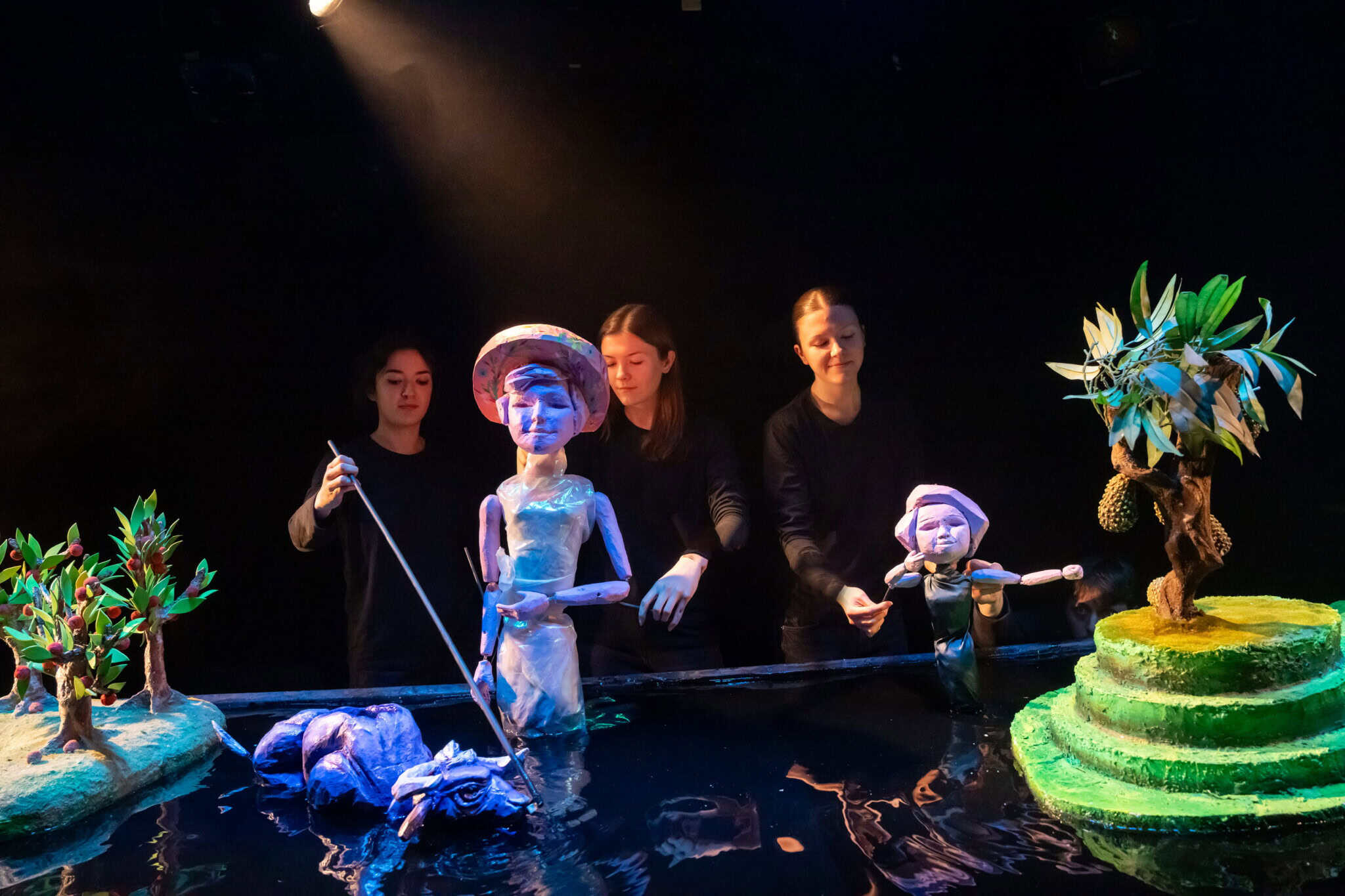 Three puppeteers dressed in black control large, colorful puppets of people, trees, and a small boat on a dark stage under dramatic lighting. The scene has a whimsical, theatrical atmosphere.