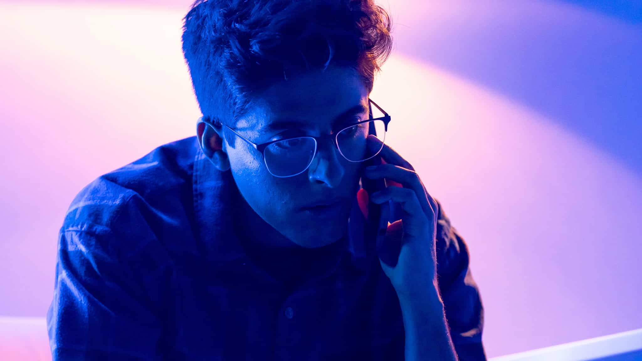 A person wearing glasses and a plaid shirt is sitting in dramatic blue and purple lighting, holding a phone to their ear and looking intently ahead, as if analyzing Karan Brar data.