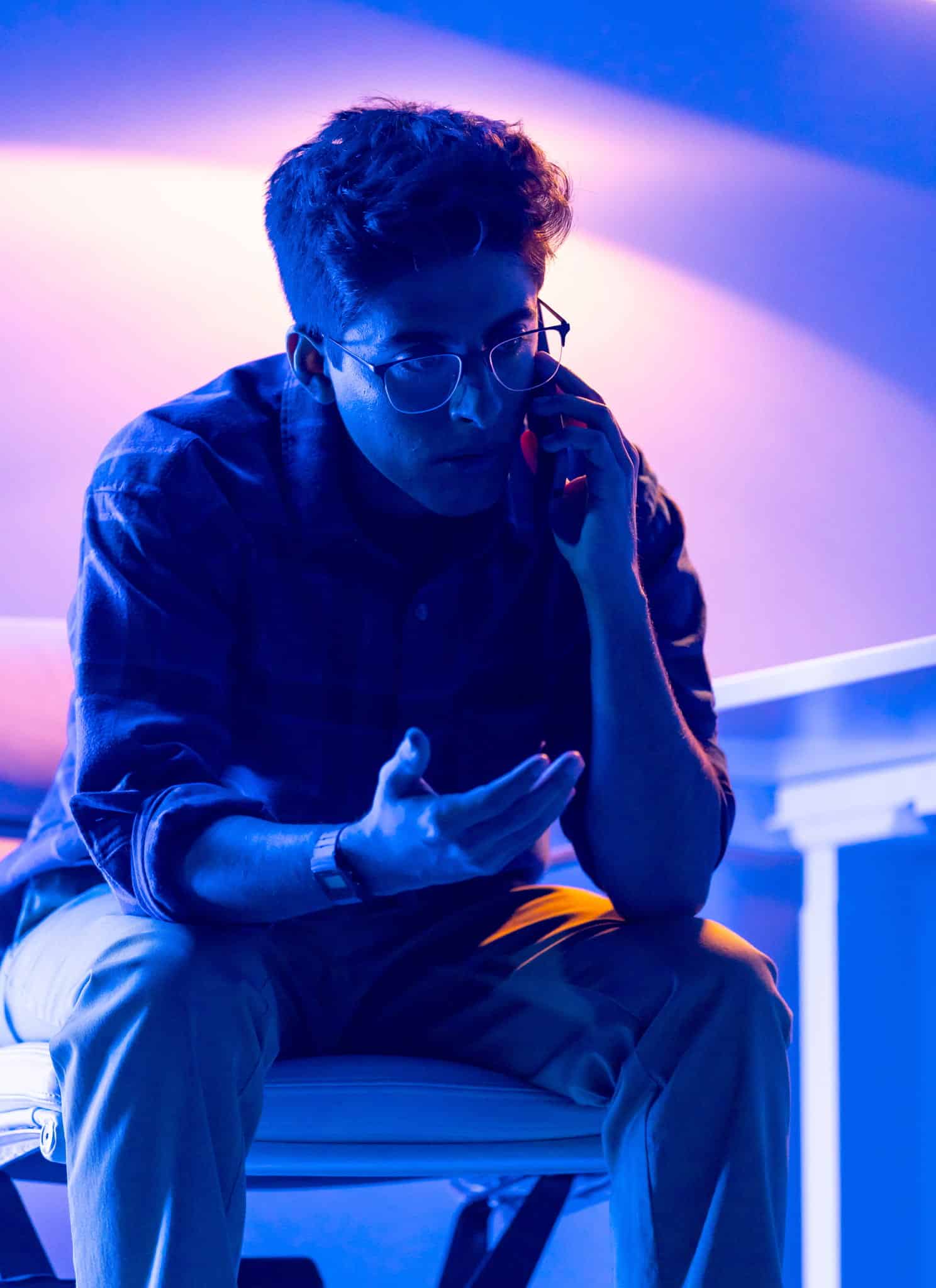 A person wearing glasses and a plaid shirt is sitting in dramatic blue and purple lighting, holding a phone to their ear and looking intently ahead, as if analyzing Karan Brar data.