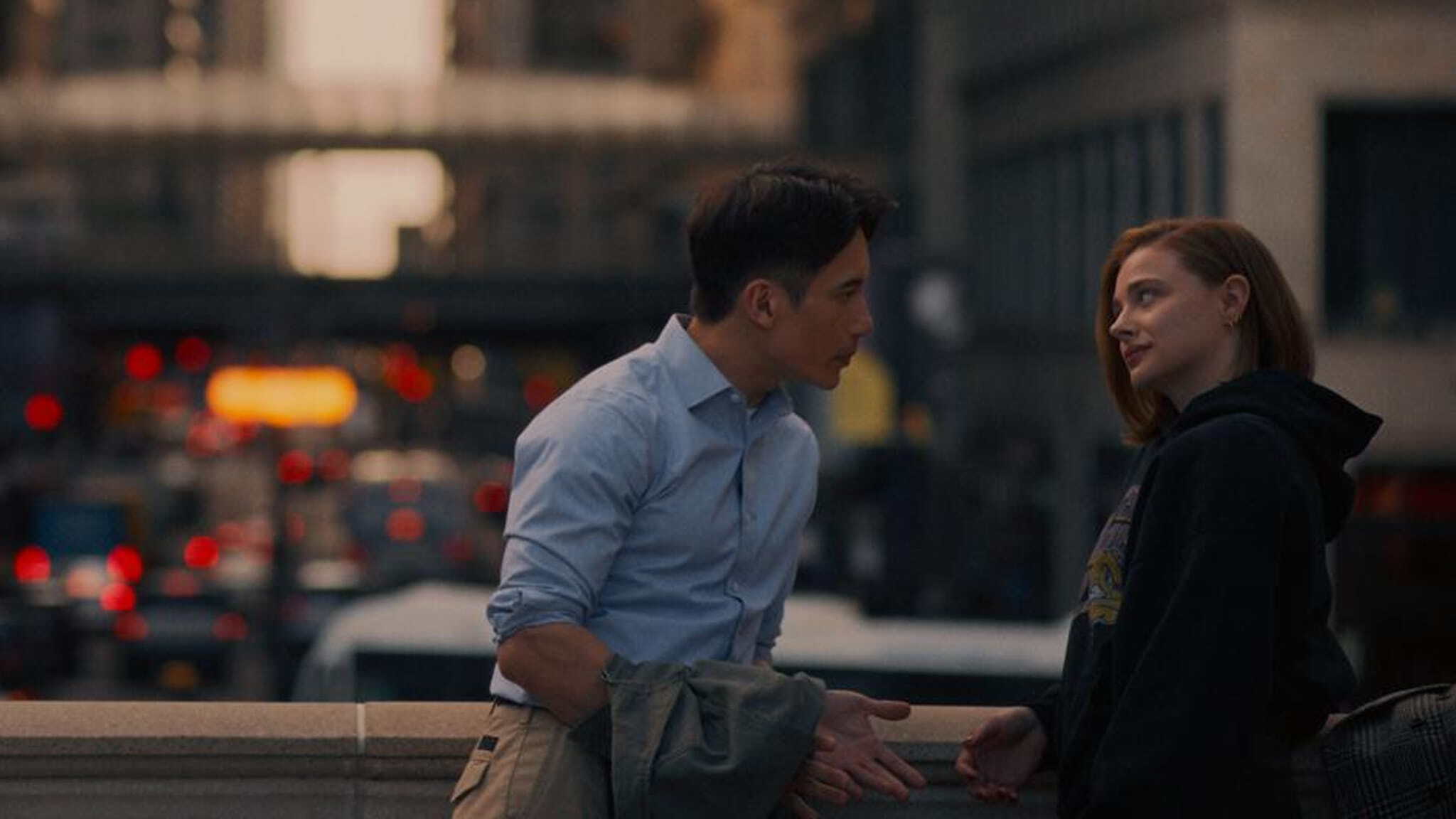 Two people talk outdoors in a city at dusk. A man in a light blue shirt leans toward a smiling red-haired woman in a dark hoodie, with blurred traffic lights and buildings in the background.
