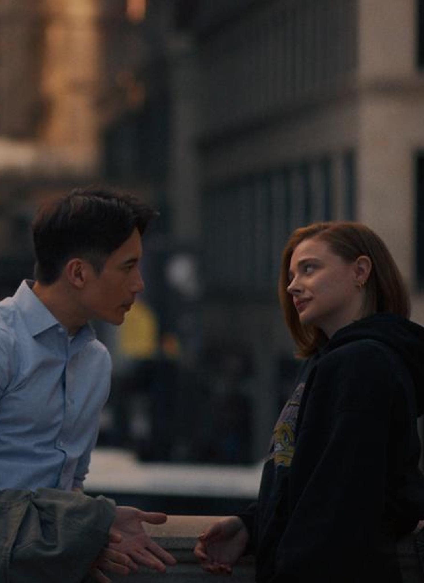 Two people talk outdoors in a city at dusk. A man in a light blue shirt leans toward a smiling red-haired woman in a dark hoodie, with blurred traffic lights and buildings in the background.