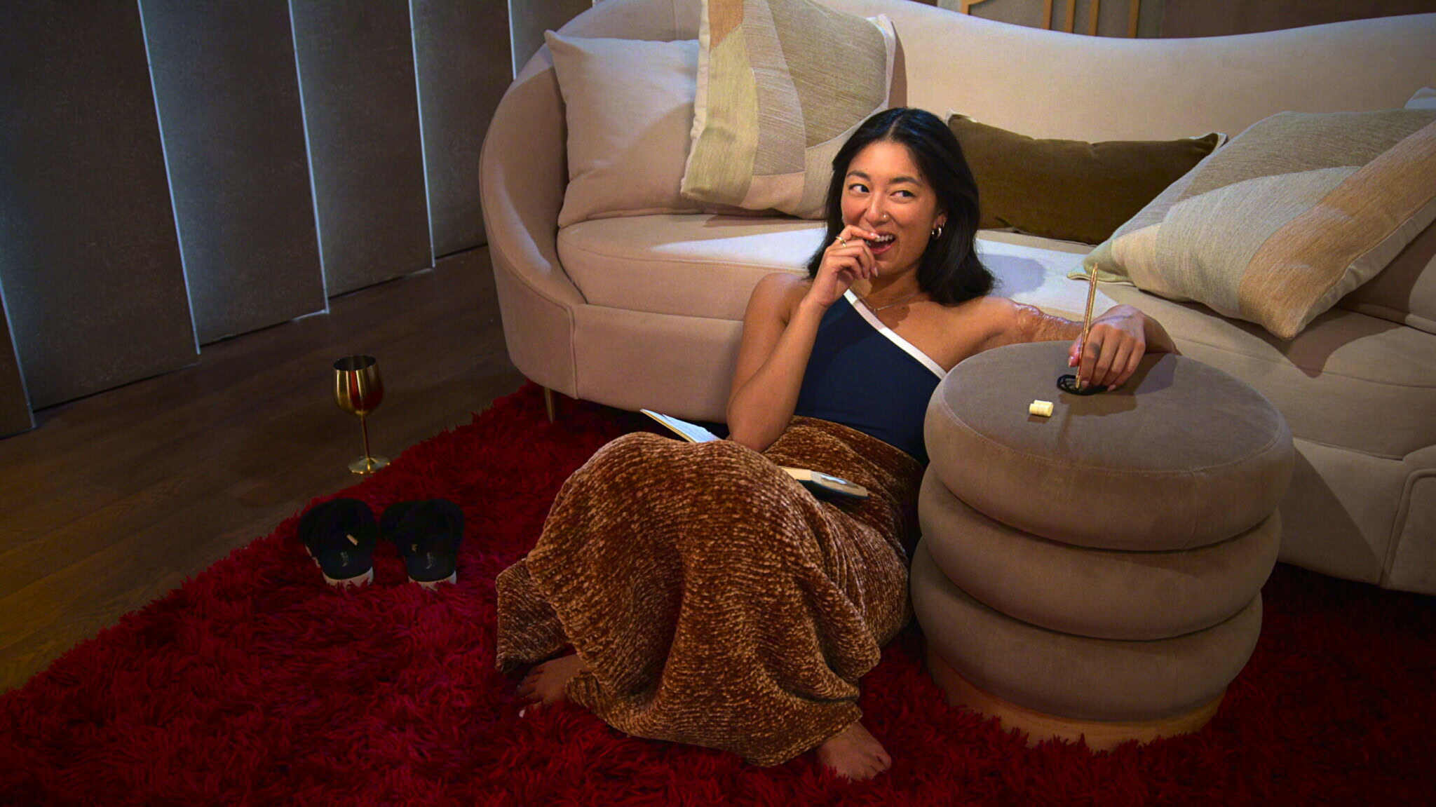 A woman sits on a red rug beside a beige couch, wrapped in a brown blanket, holding a snack and smiling. There’s a notebook, slippers, and a glass of wine nearby, creating a cozy, relaxed atmosphere.