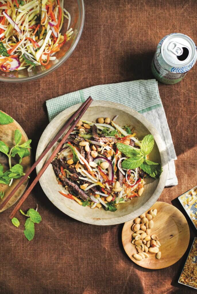 A bowl of noodle salad topped with grilled meat, sliced vegetables, peanuts, and mint, with chopsticks resting on the bowl. Nearby are a small dish of peanuts, a can of drink, and a bowl of salad.
