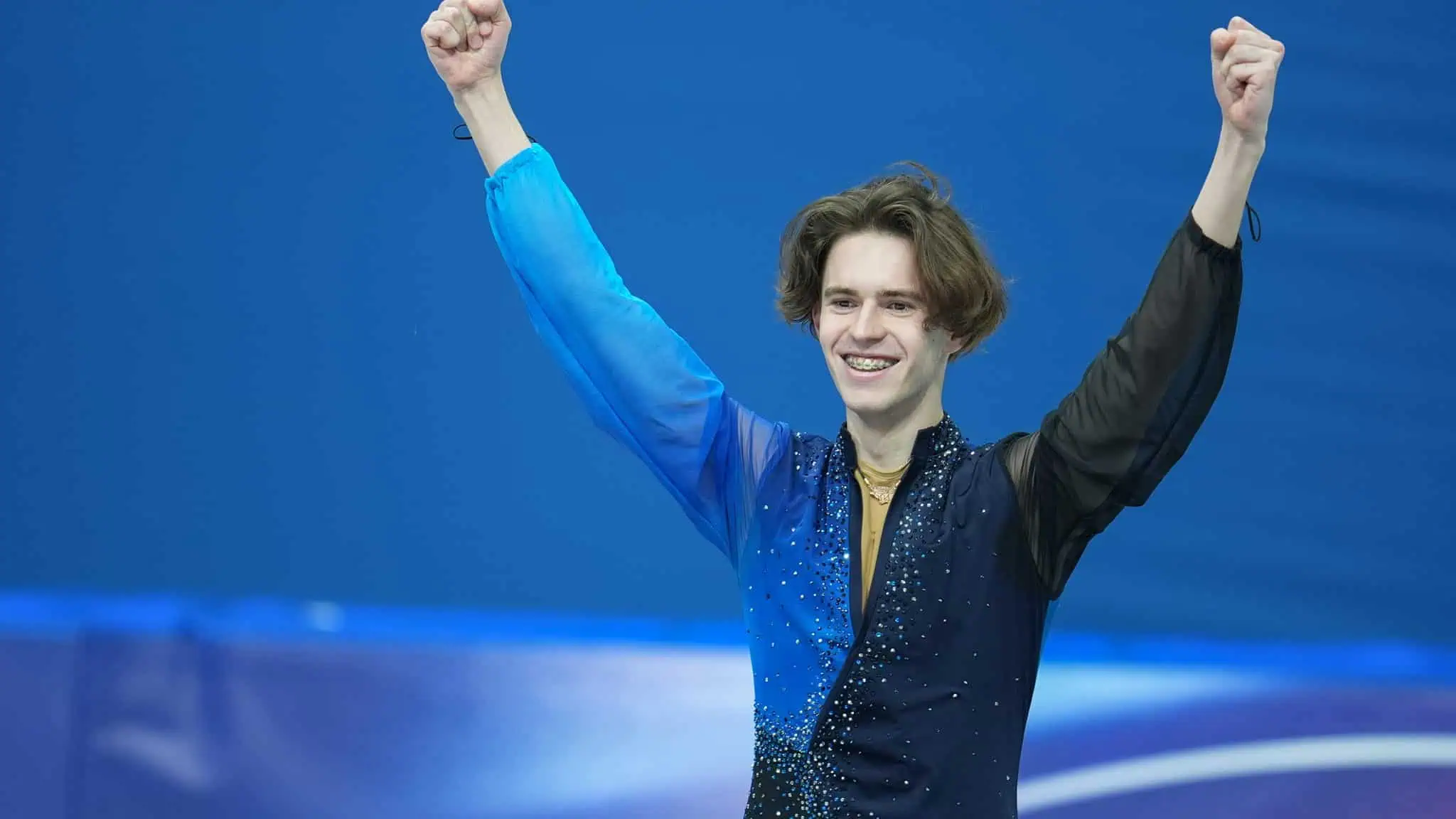 A male figure skater wearing a blue and black costume smiles and raises both arms in celebration on the ice rink, with a blue background behind him.