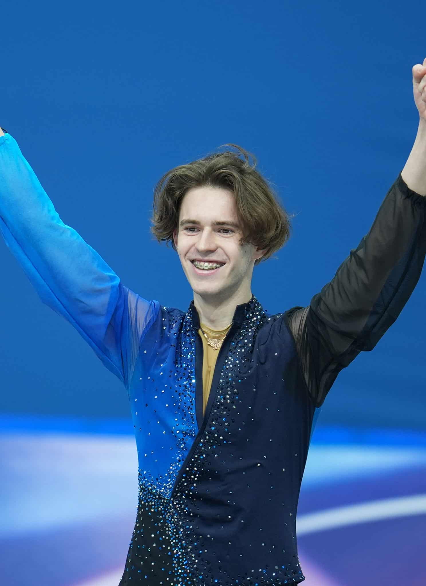 A male figure skater wearing a blue and black costume smiles and raises both arms in celebration on the ice rink, with a blue background behind him.