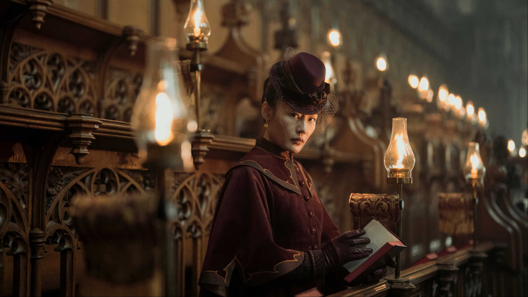 A woman dressed in Victorian-era clothing sits in a dimly lit church pew, holding a book and gazing intently ahead. Oil lamps line the ornate wooden pews, casting a warm glow.