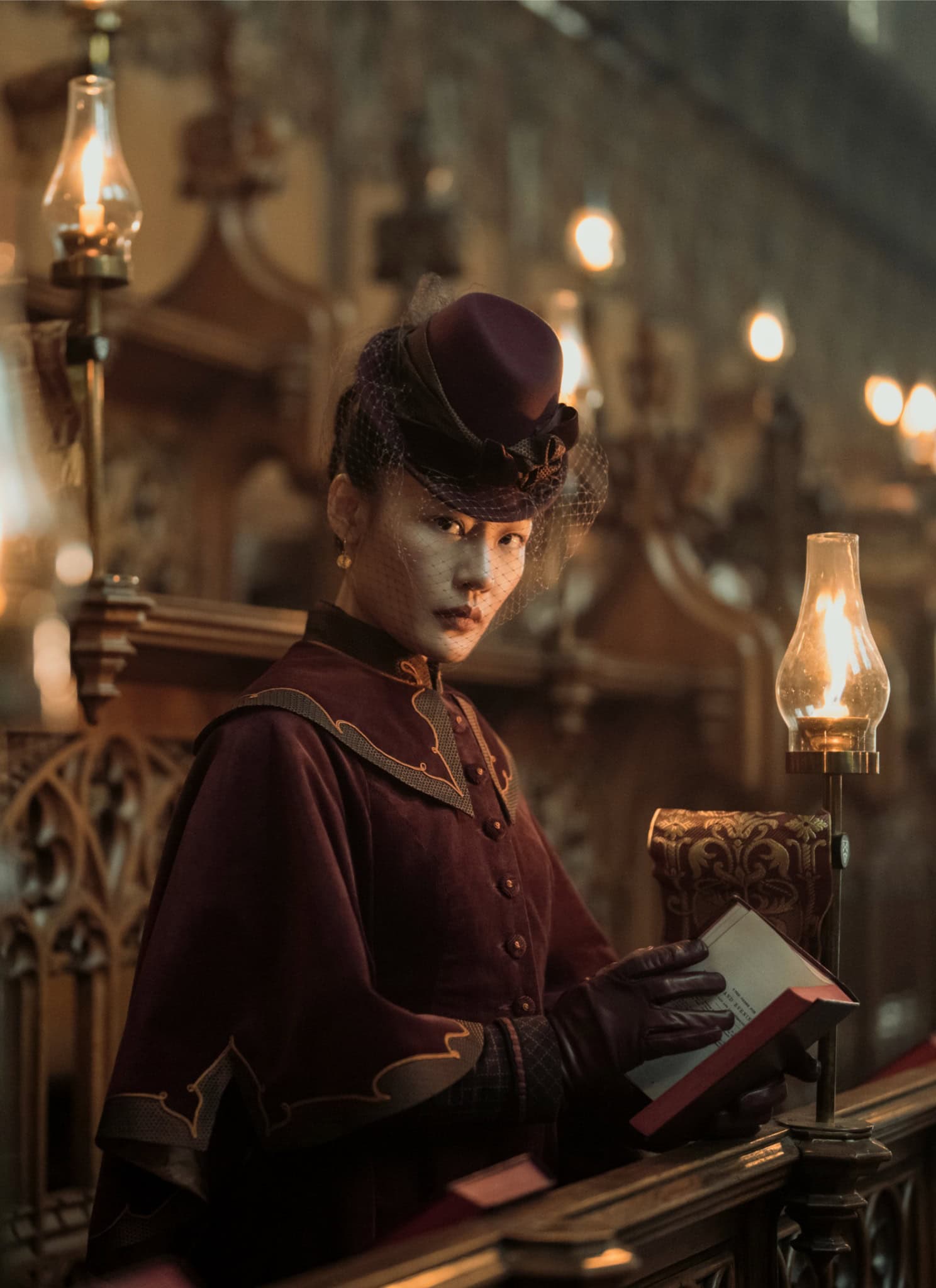 A woman dressed in Victorian-era clothing sits in a dimly lit church pew, holding a book and gazing intently ahead. Oil lamps line the ornate wooden pews, casting a warm glow.