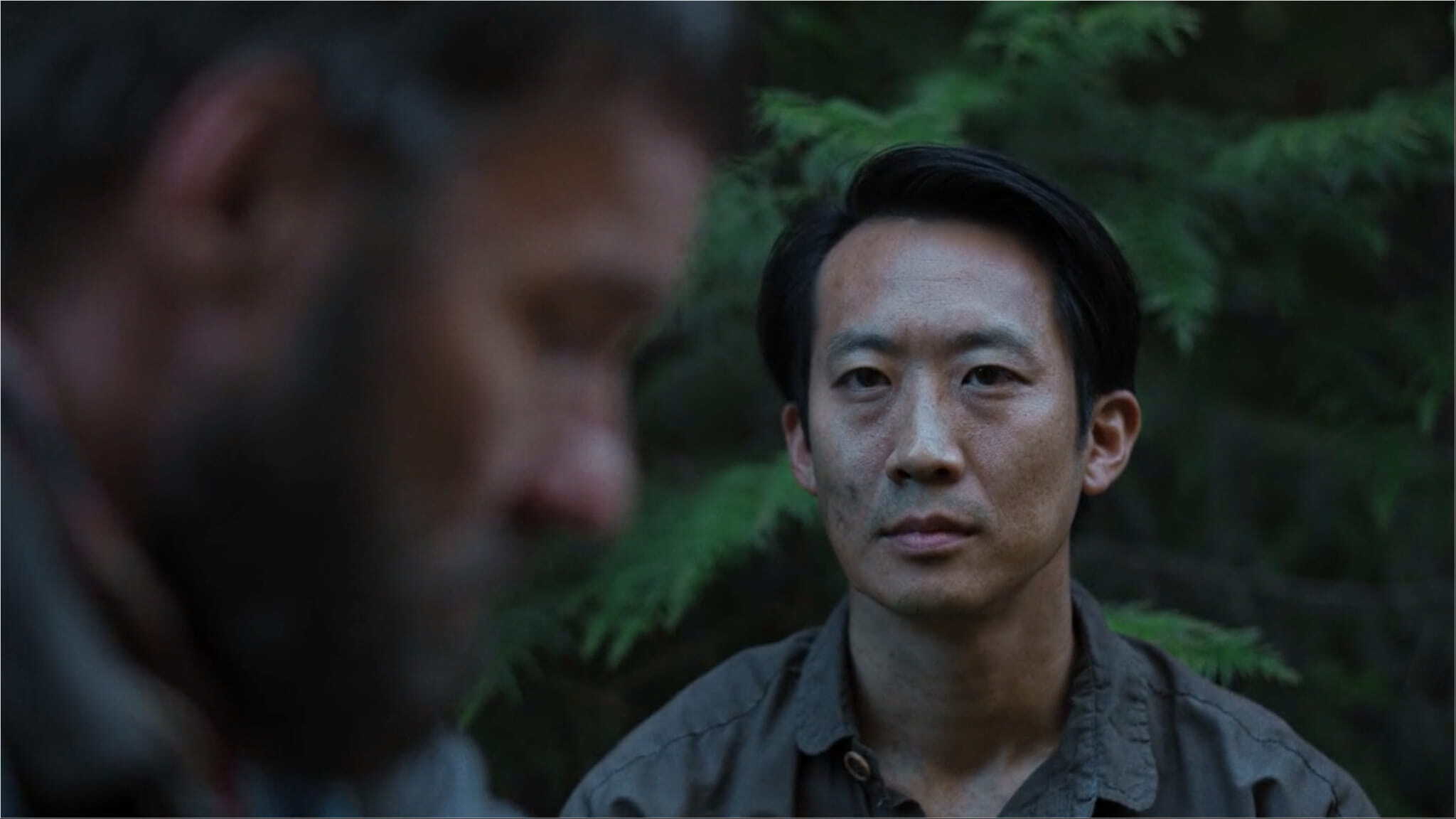 A man with short dark hair looks intently at another man in the foreground, who is out of focus. They are outdoors, with green foliage in the background.