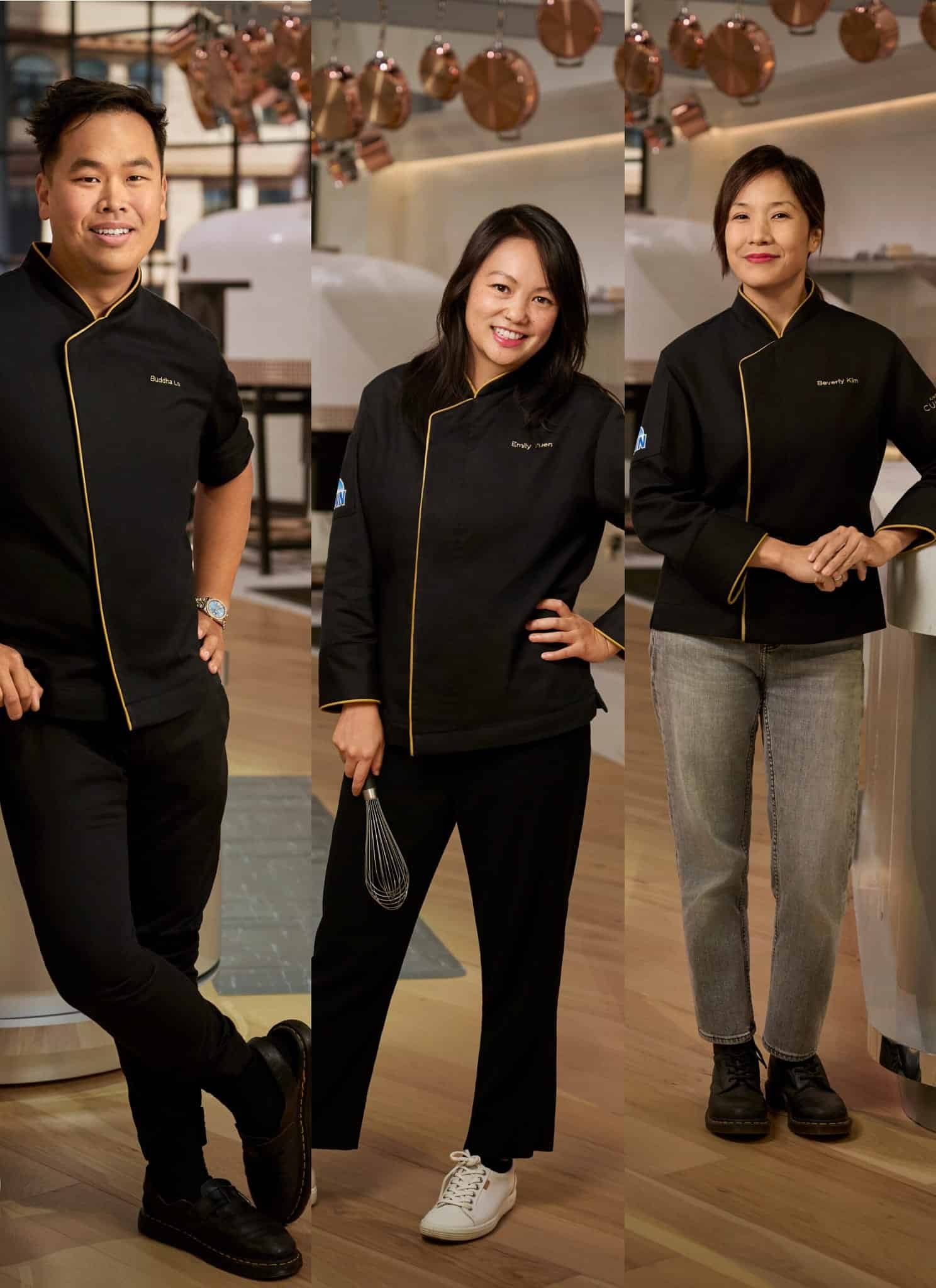 Three chefs in black uniforms pose in a modern, brightly lit kitchen. The kitchen features wooden floors, white counters, hanging copper pans, and large ovens in the background. Each chef stands confidently and smiles.