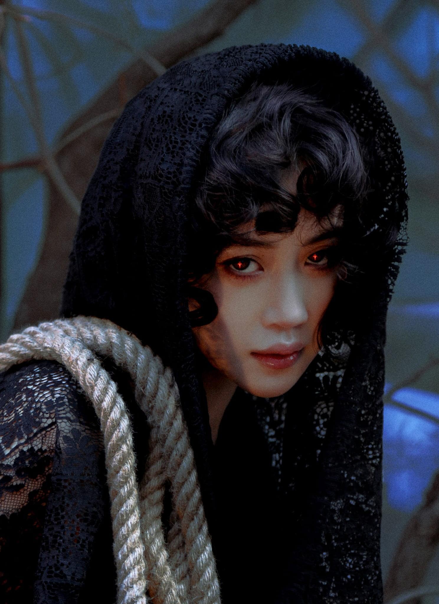 A person with pale skin, curly dark hair, and glowing red eyes gazes intensely. Wearing a black lace hooded cloak, they have a thick rope over one shoulder. The dark, blurred background with bare branches sets a mysterious DeVita-like tone.