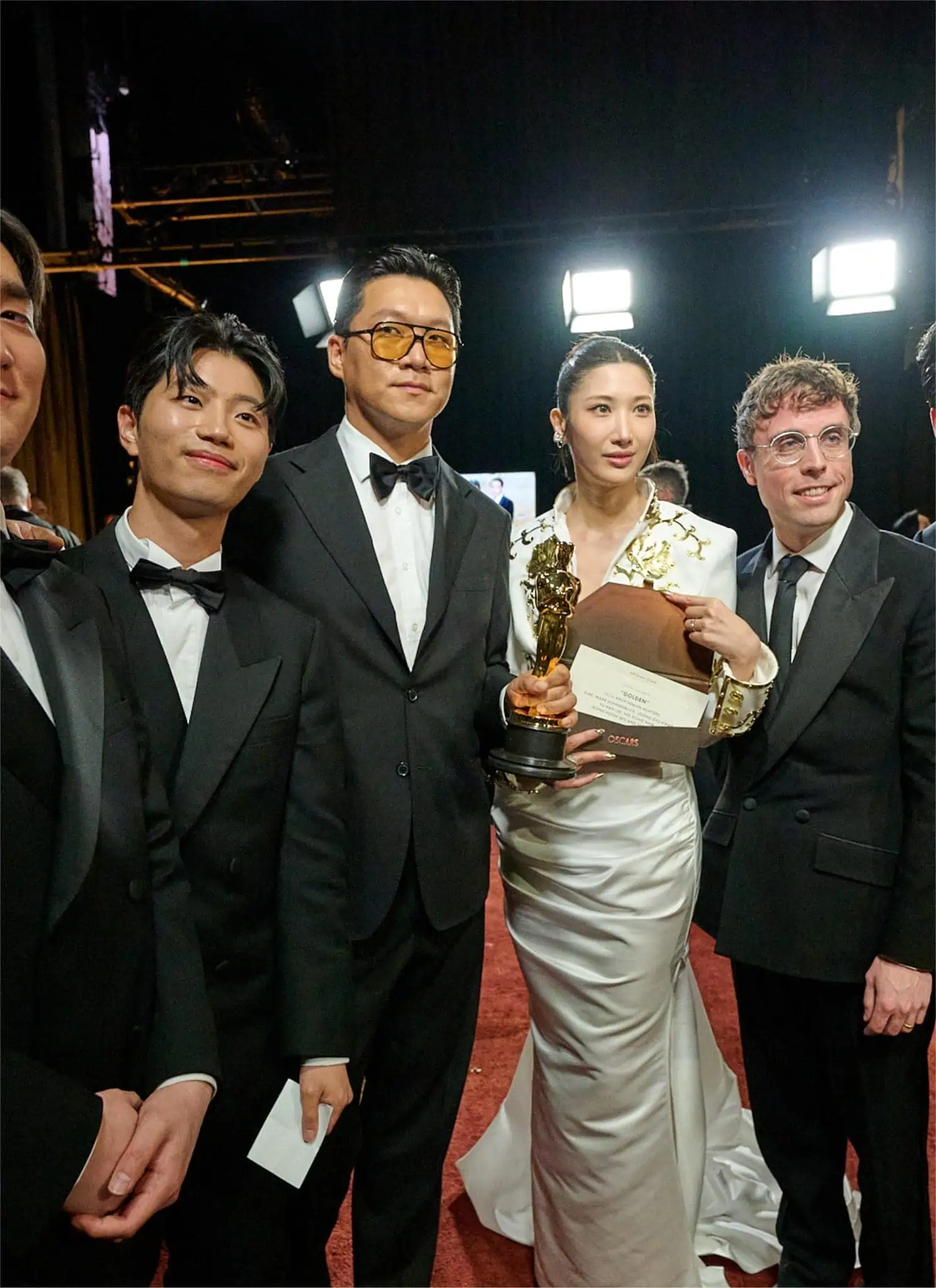 A group of five men in black tuxedos and one woman in a white gown stand together smiling at an event; the woman is holding a gold trophy and an envelope.