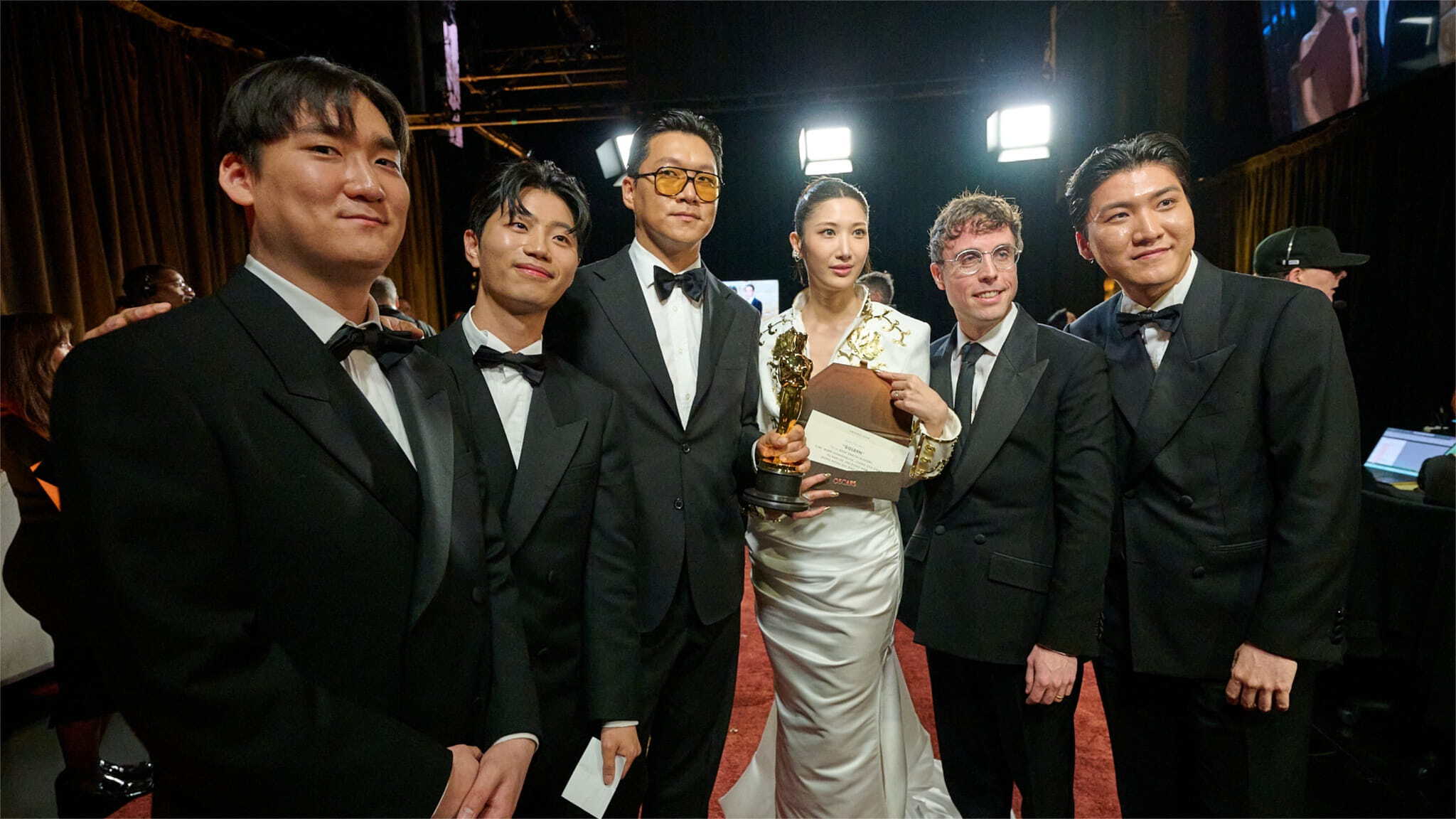 A group of five men in black tuxedos and one woman in a white gown stand together smiling at an event; the woman is holding a gold trophy and an envelope.