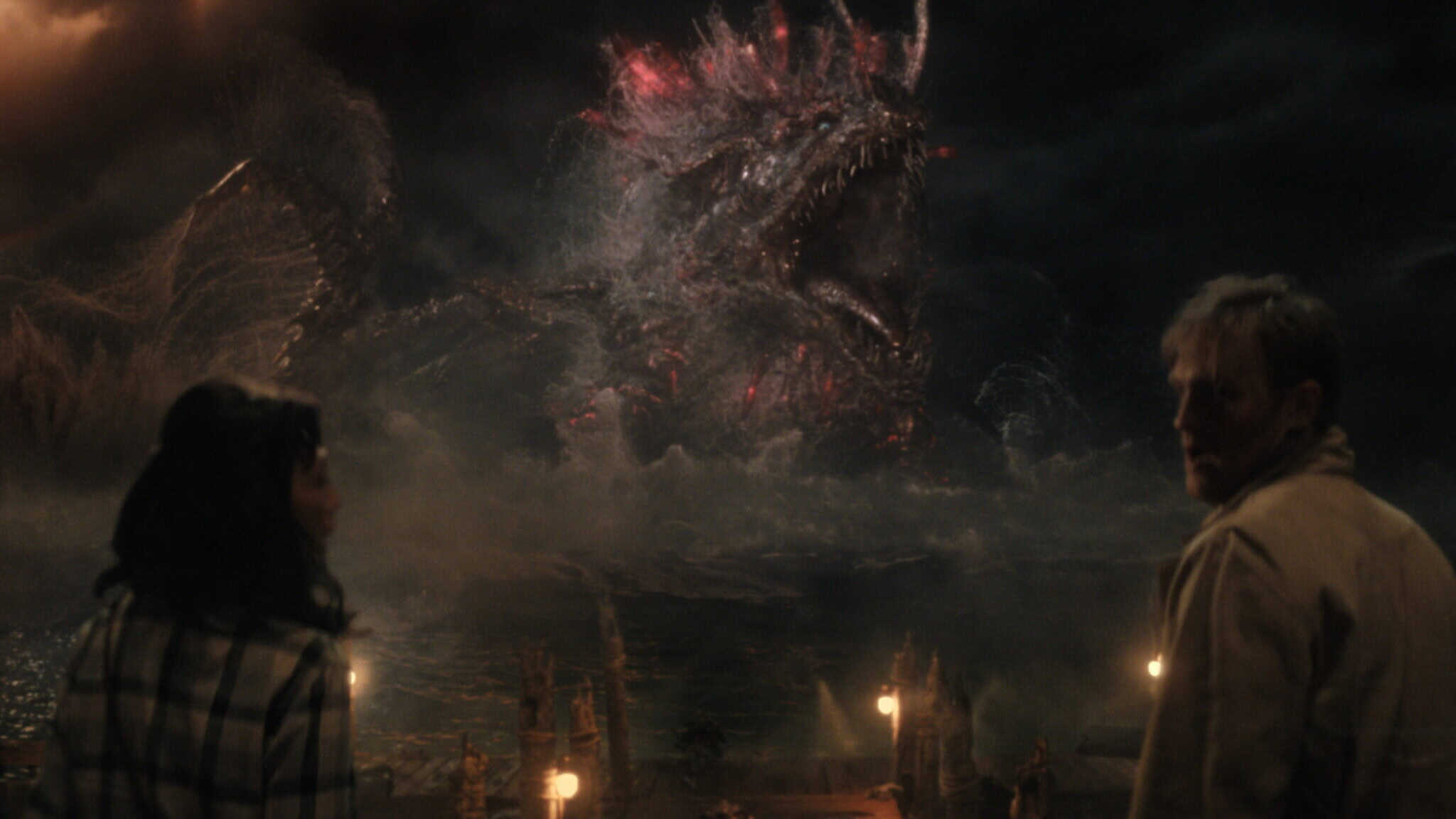 Two people stand on a pier at night, gazing toward a giant, glowing sea monster—much like a creature from Monarch: Legacy of Monsters—emerging from stormy waters, its mouth open wide and red light emanating from its body.