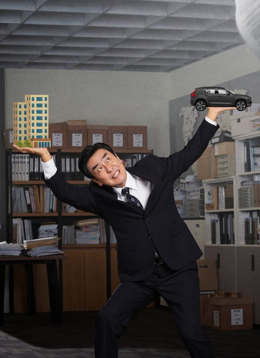 A man in a suit stands in a messy office, juggling a toy car and a model building while papers fly through the air, conveying chaos and multitasking.