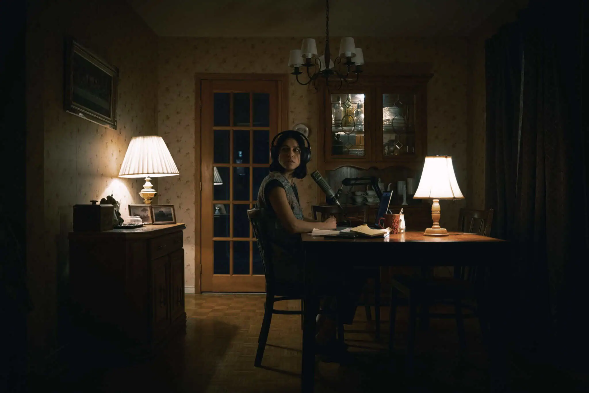 A person sits at a dimly lit dining table at night, wearing headphones and speaking into a microphone. Two table lamps and a chandelier partially illuminate the room, which has warm, cozy decor and wooden furniture.