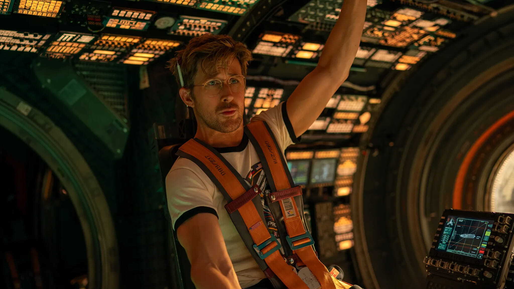 A man wearing glasses and an orange harness sits in a high-tech spacecraft cockpit, surrounded by illuminated control panels and screens, looking focused as he holds onto an overhead handle.