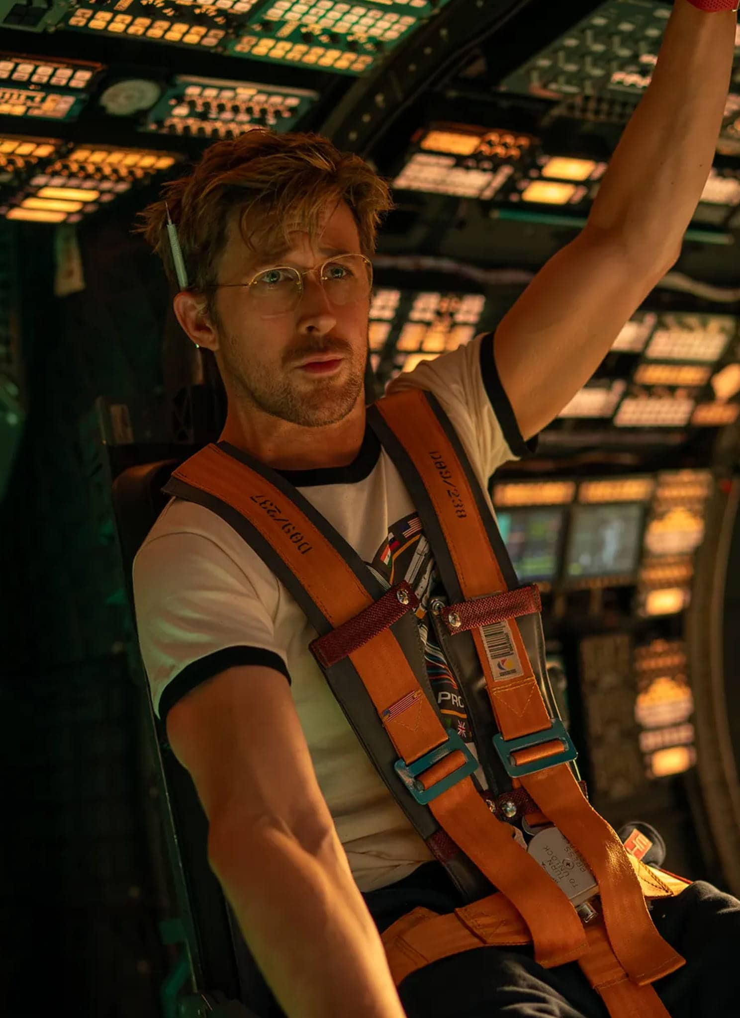 A man wearing glasses and an orange harness sits in a high-tech spacecraft cockpit, surrounded by illuminated control panels and screens, looking focused as he holds onto an overhead handle.